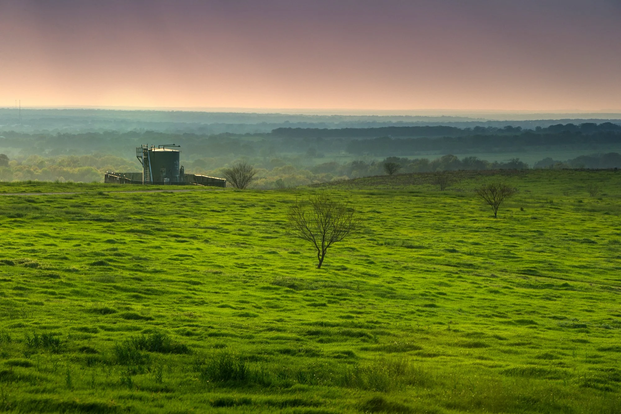 A lush green field with a few scattered trees, hills in the distance, and a foggy sky at sunset, with a large silos structure on the left side of the image.