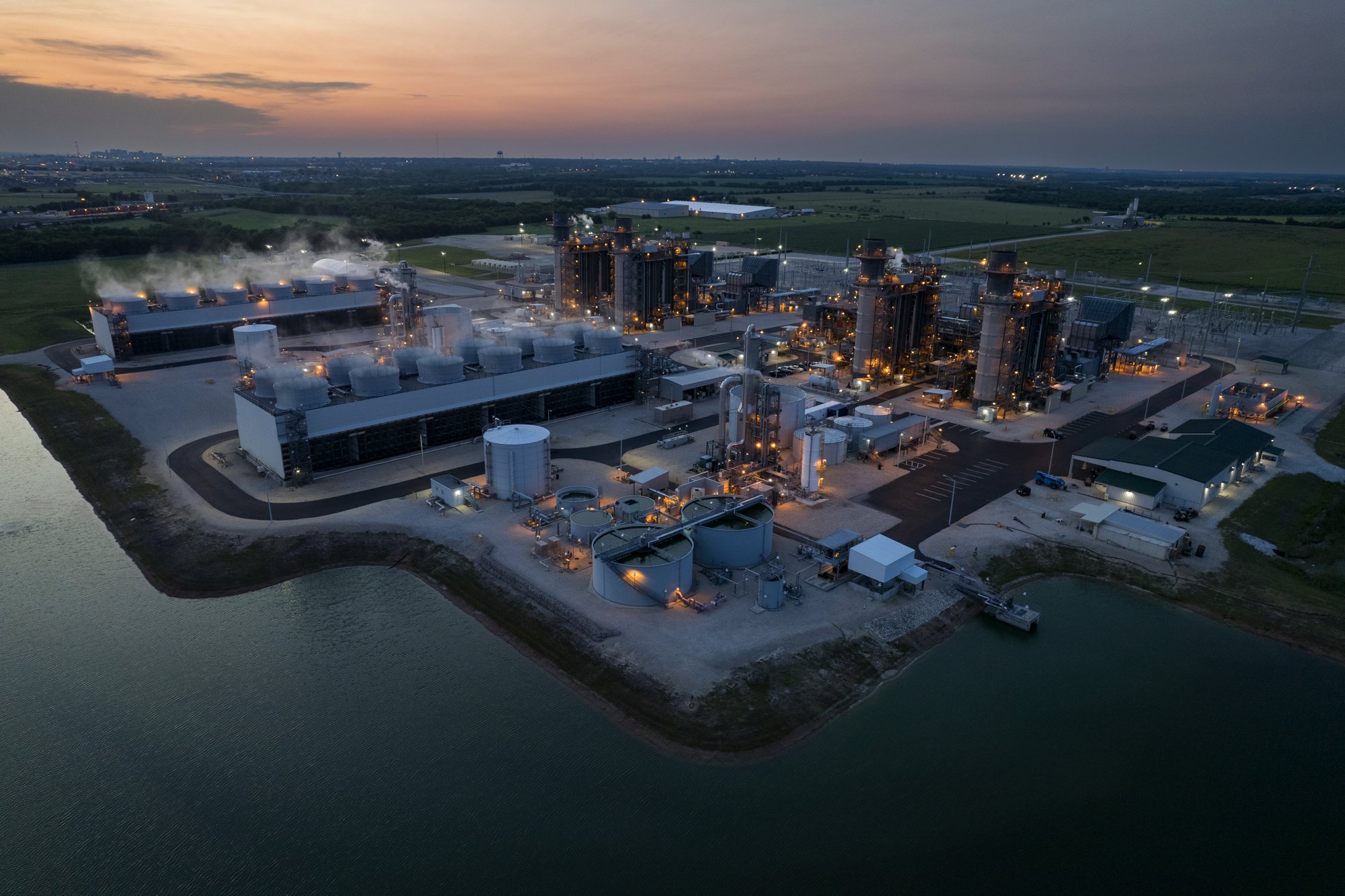 Aerial view of an industrial complex near a body of water at dusk, with multiple buildings, storage tanks, and smokestacks emitting steam or smoke.