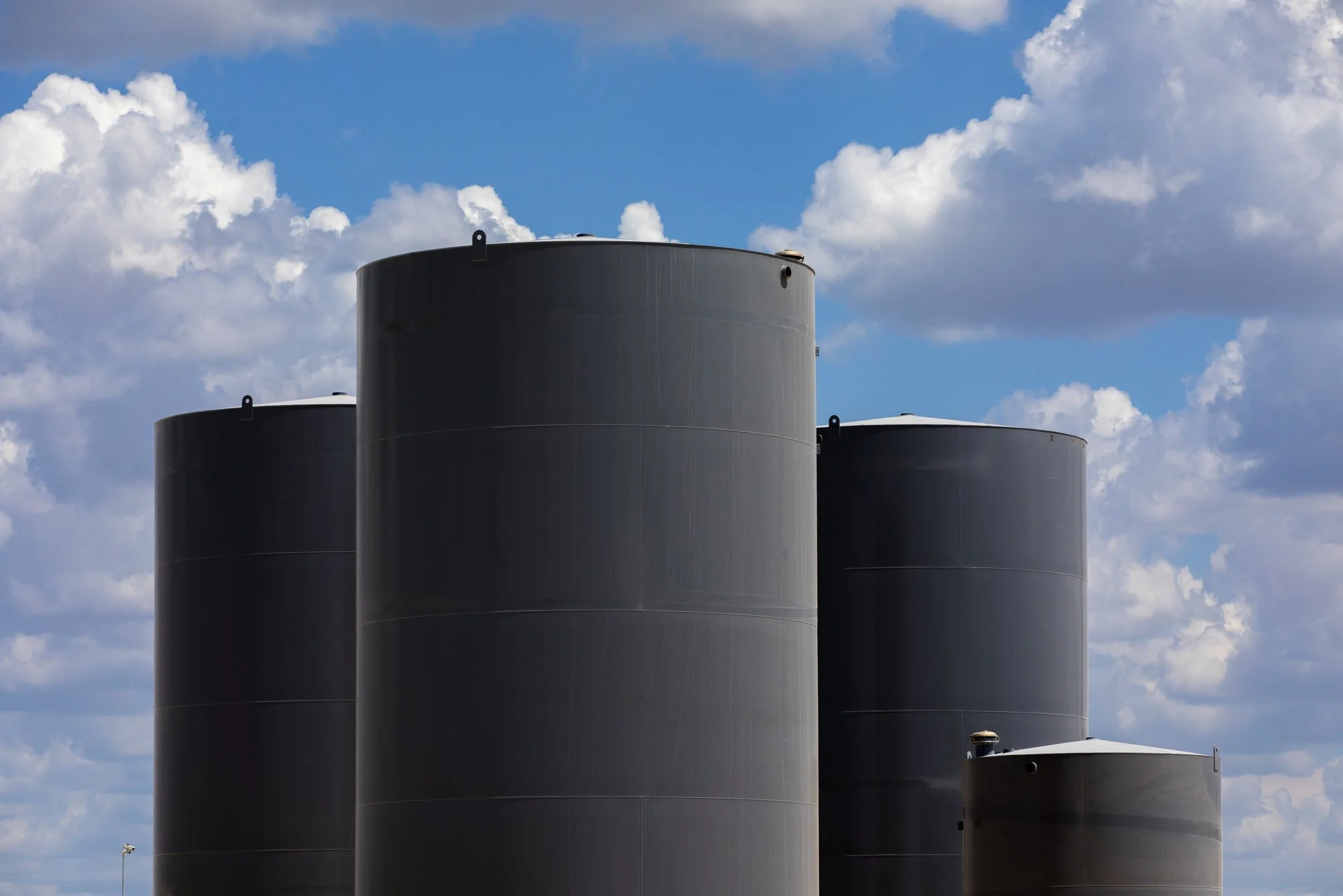 Four large black industrial storage tanks against a cloudy blue sky.