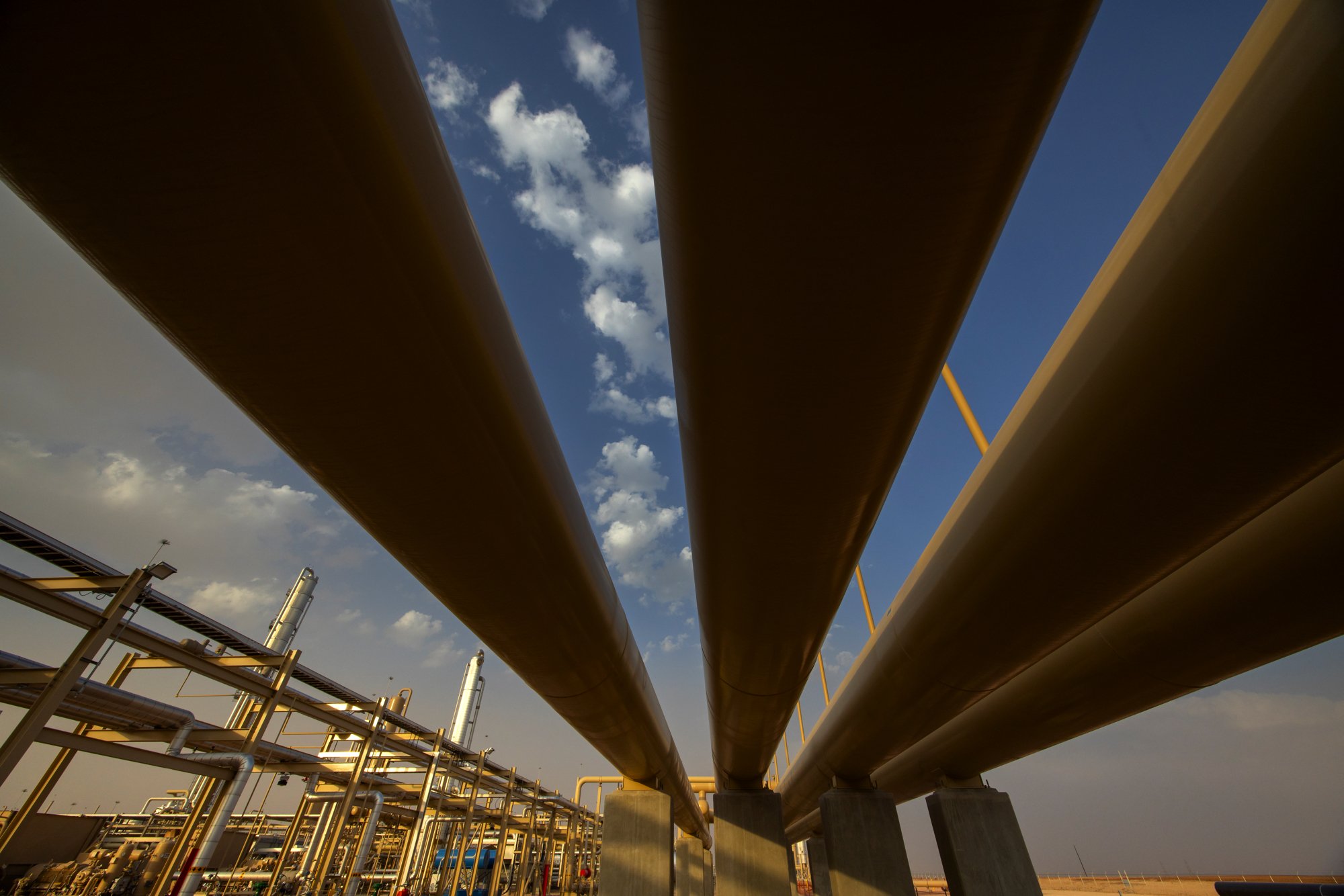 Large industrial pipes running parallel above an industrial facility with pipes and structures, against a partly cloudy sky.