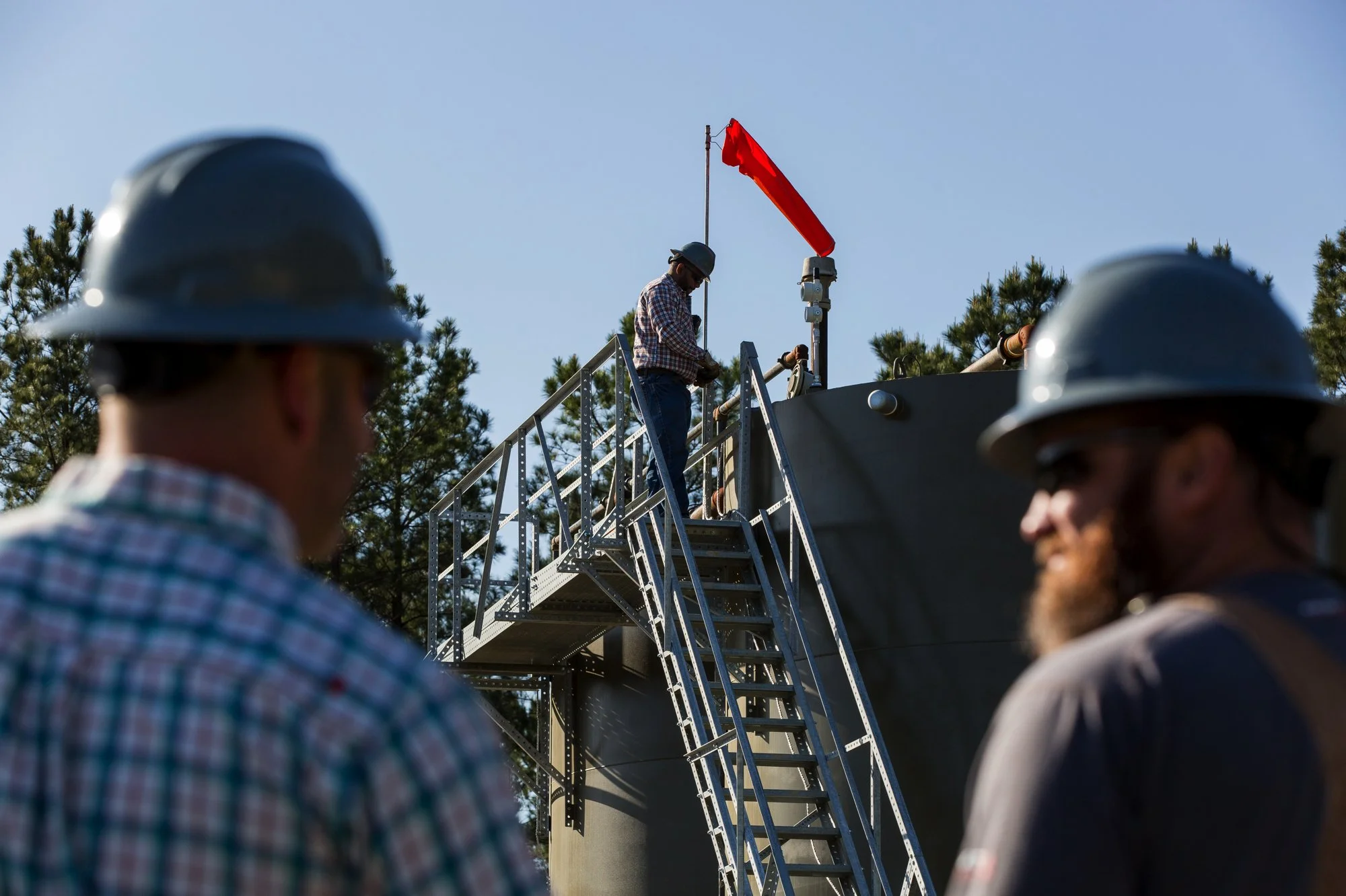 Workers with safety helmets inspecting a metal structure with a ladder and a person working on top of the structure, outdoors with trees and a clear sky.
