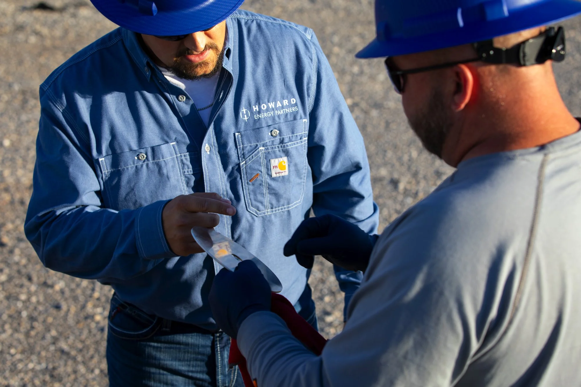 Two workers outdoors in safety helmets and gloves exchanging a metal plate or sample, with gravel ground visible.