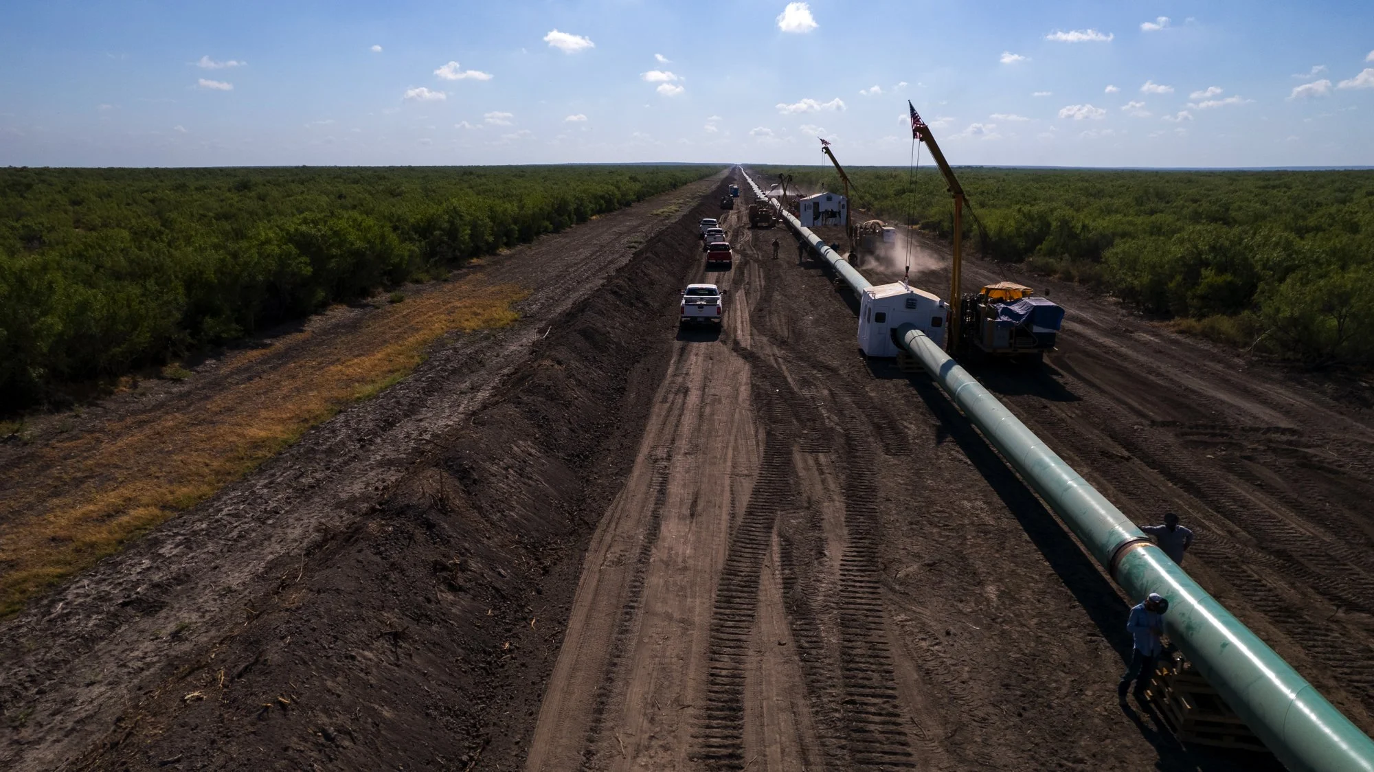 Construction crews work on installing a large pipeline along a dirt road in a rural area with green trees on both sides, under a partly cloudy sky.