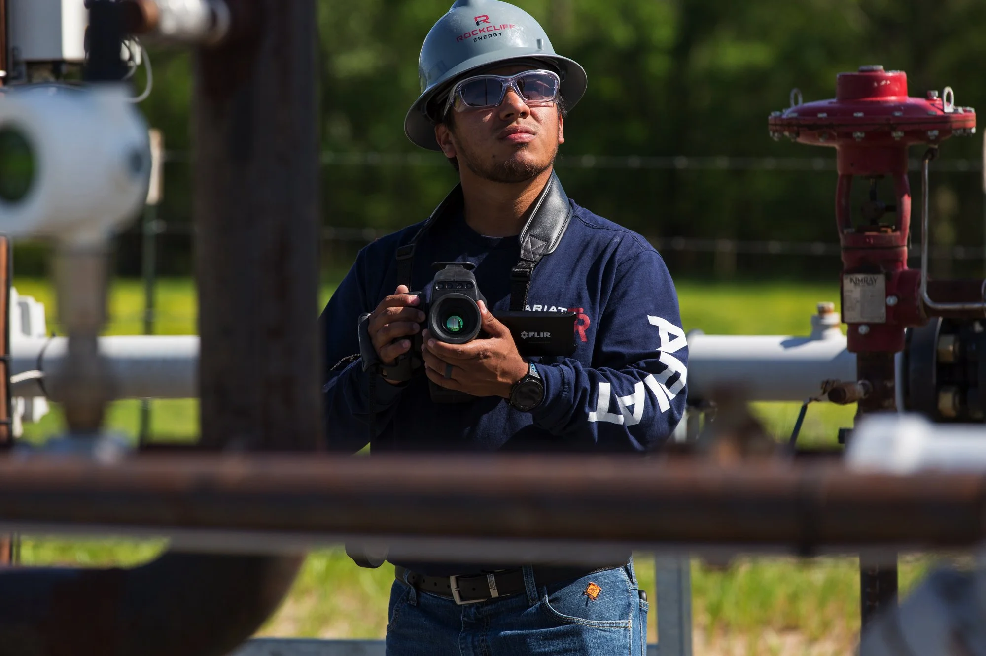A worker wearing a safety helmet, sunglasses, and a navy blue jacket with logos, holding a camera, standing outdoors near industrial pipelines and equipment.