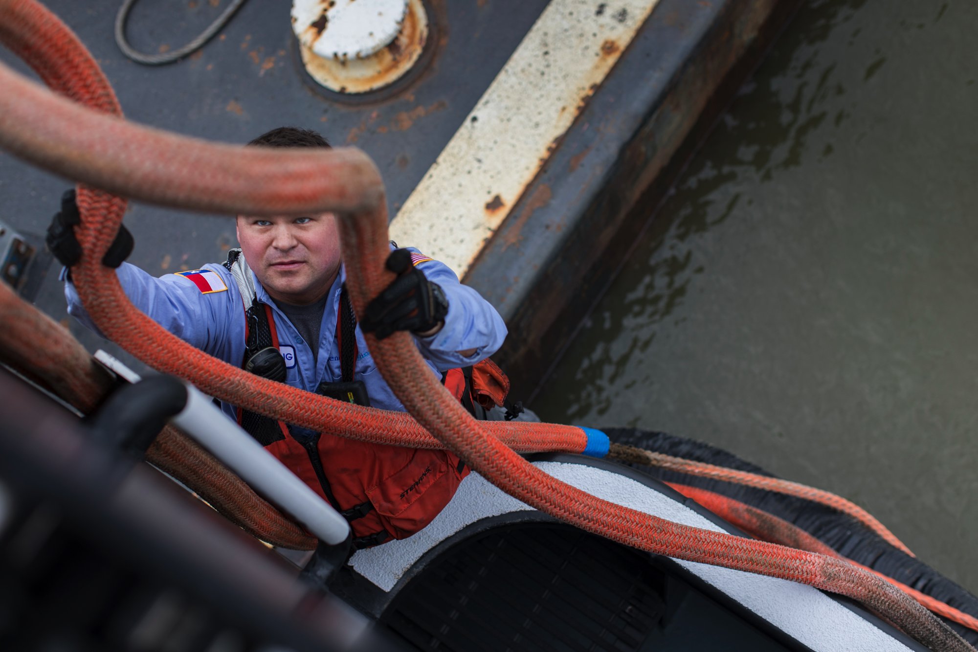 Male firefighter wearing a blue uniform and safety gear, climbing a ladder on a fire truck near water, holding onto orange ropes.