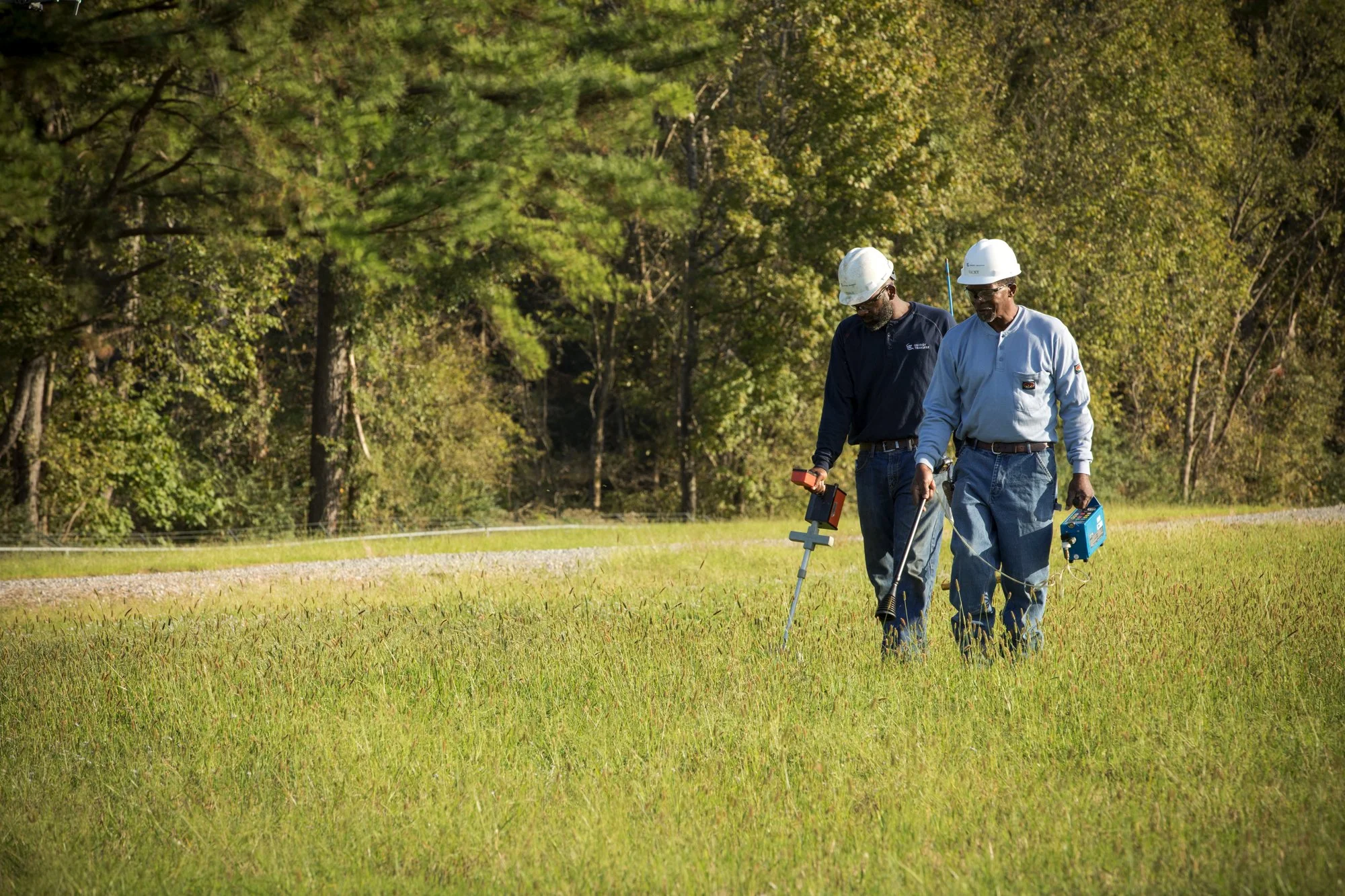 Two men wearing safety helmets and carrying surveying equipment walk through a grassy field with trees in the background.