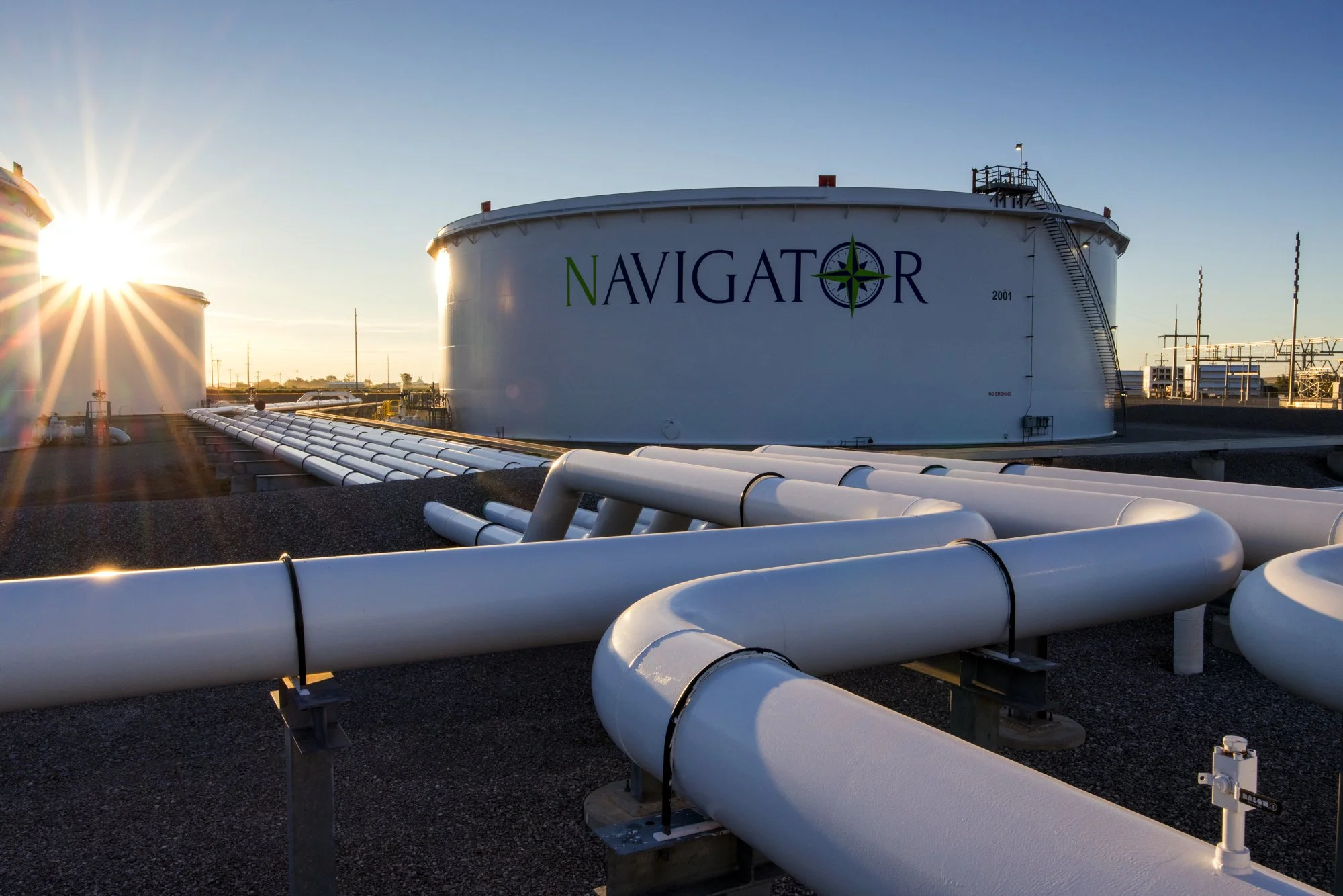 Industrial roof with large white storage tank labeled 'Navigator' and connected white pipelines, with the sun setting in the background.