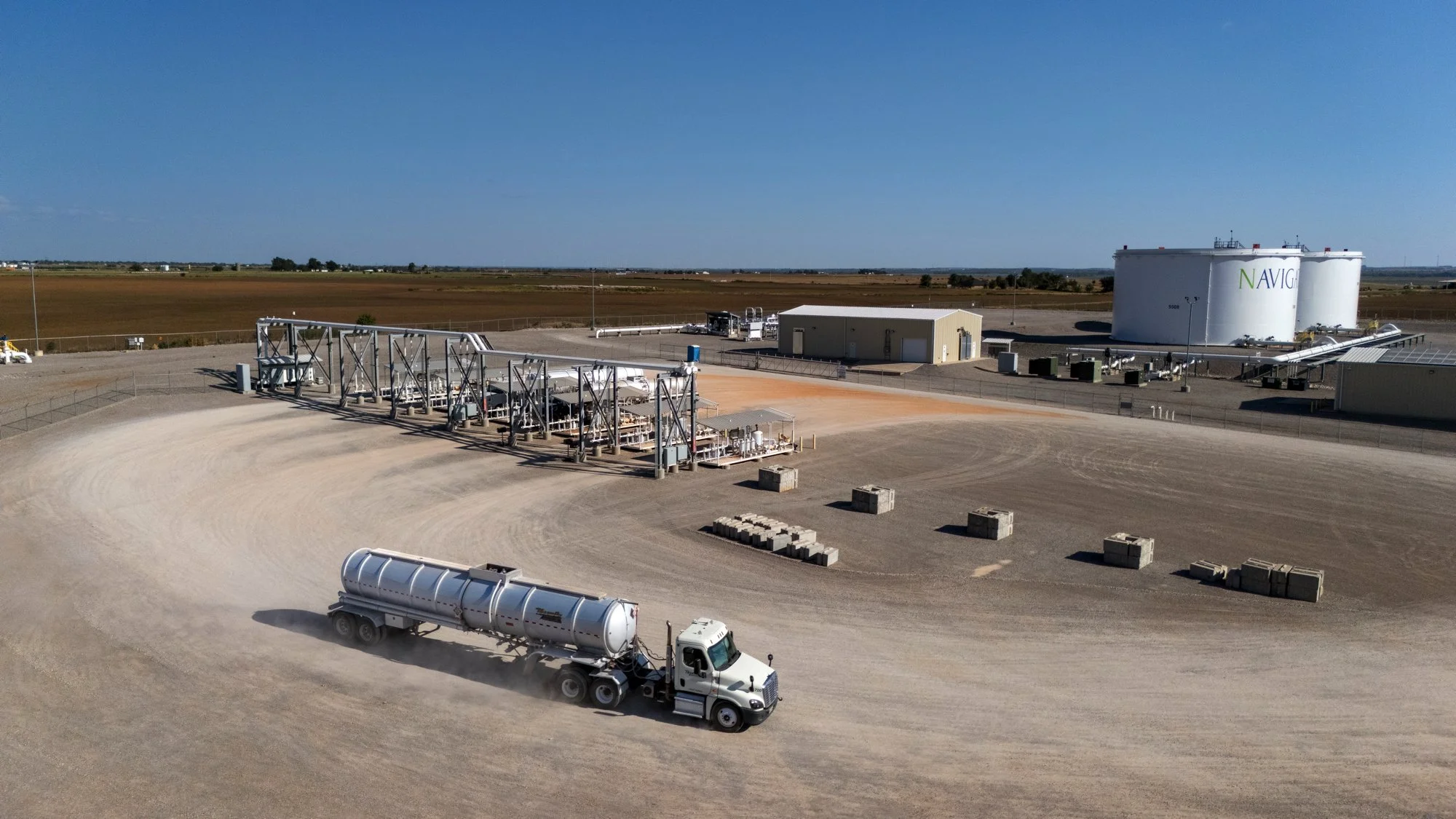 Large industrial fuel storage facility with a tanker truck driving on a gravel road in front. Multiple pipelines, storage tanks, and buildings are visible under a clear blue sky.