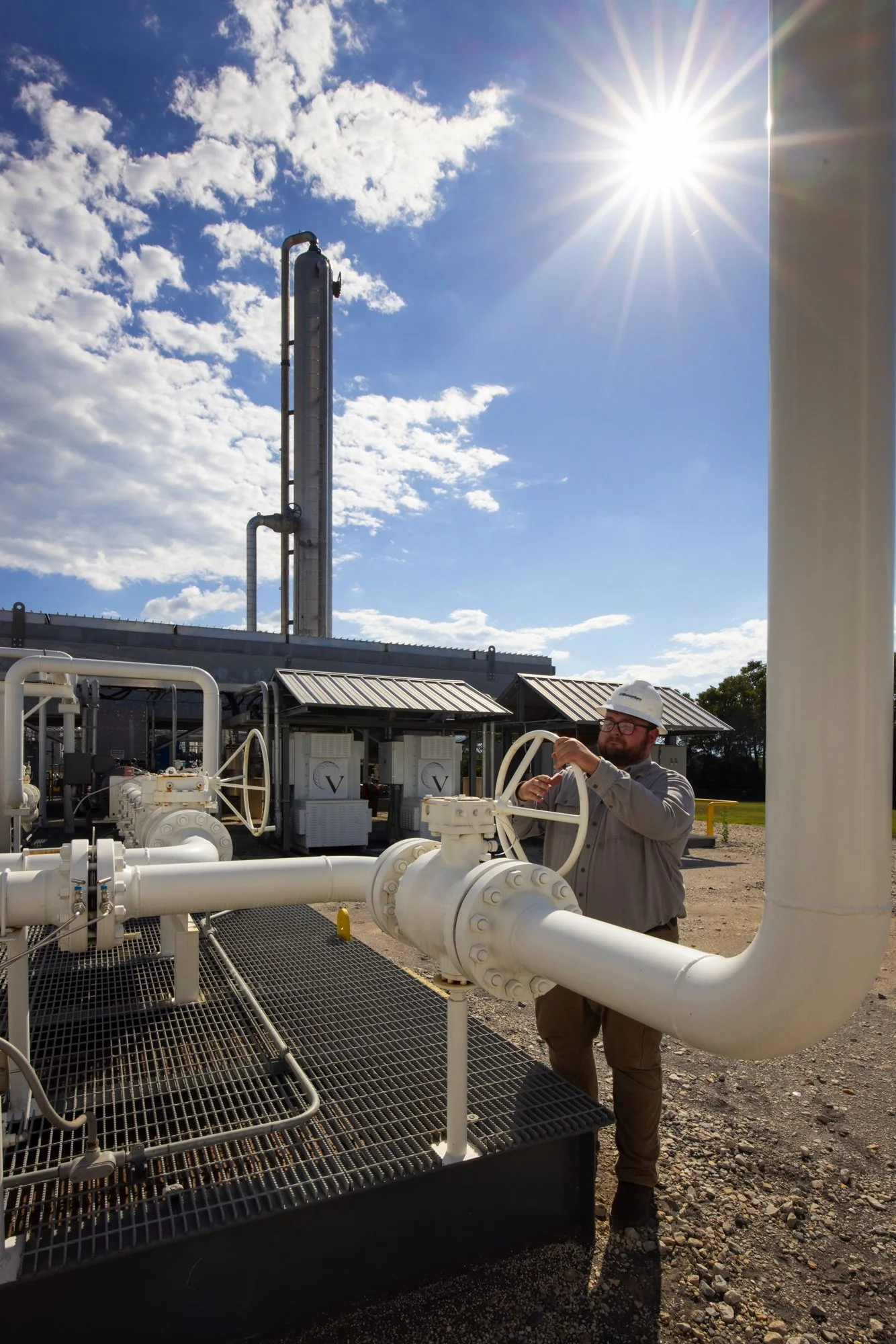 A man in safety gear operating valves at an oil or gas plant with piping and equipment under a clear blue sky with some clouds.