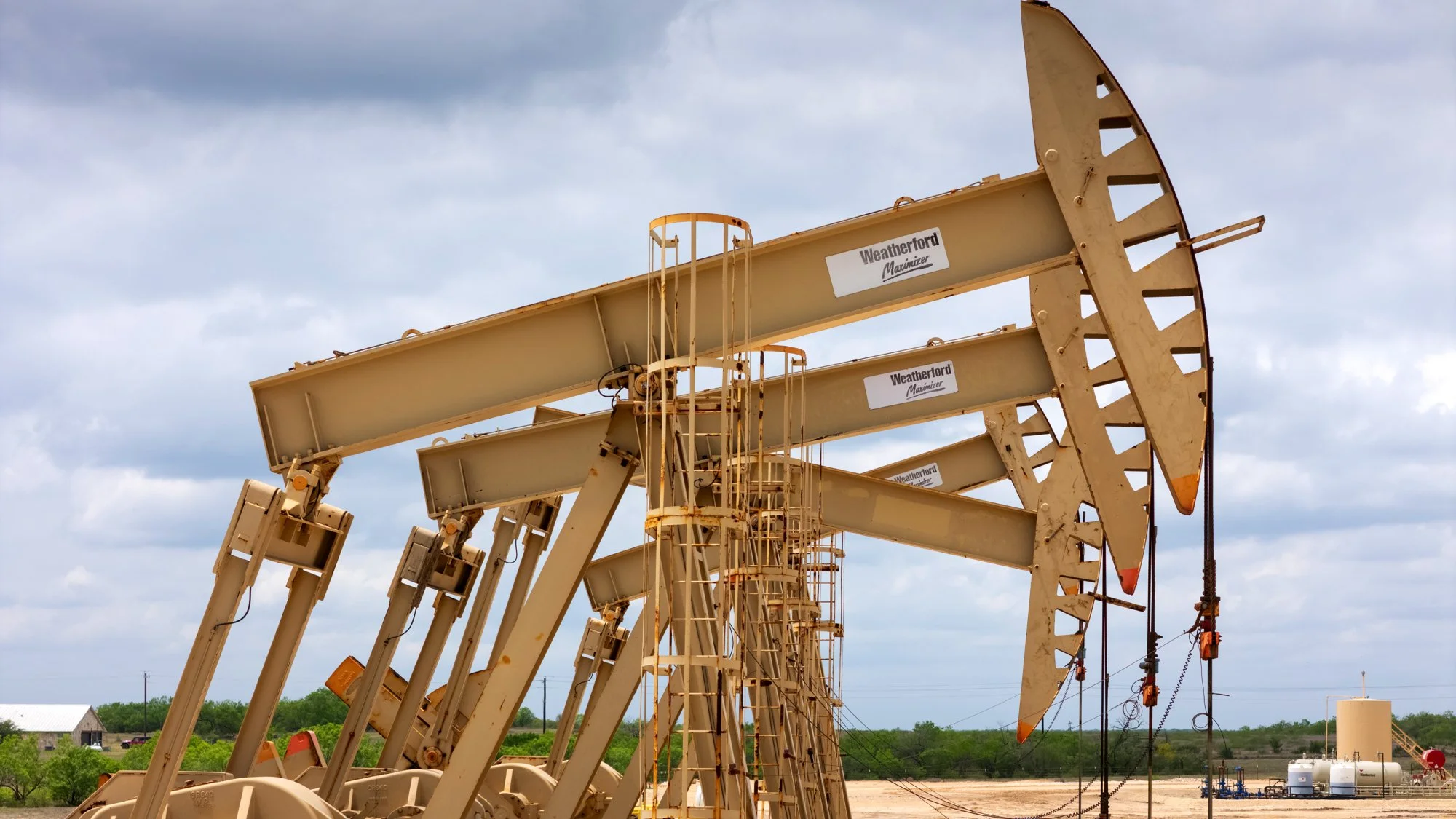 Several beige oil pump jacks working in an open oil field under a partly cloudy sky.