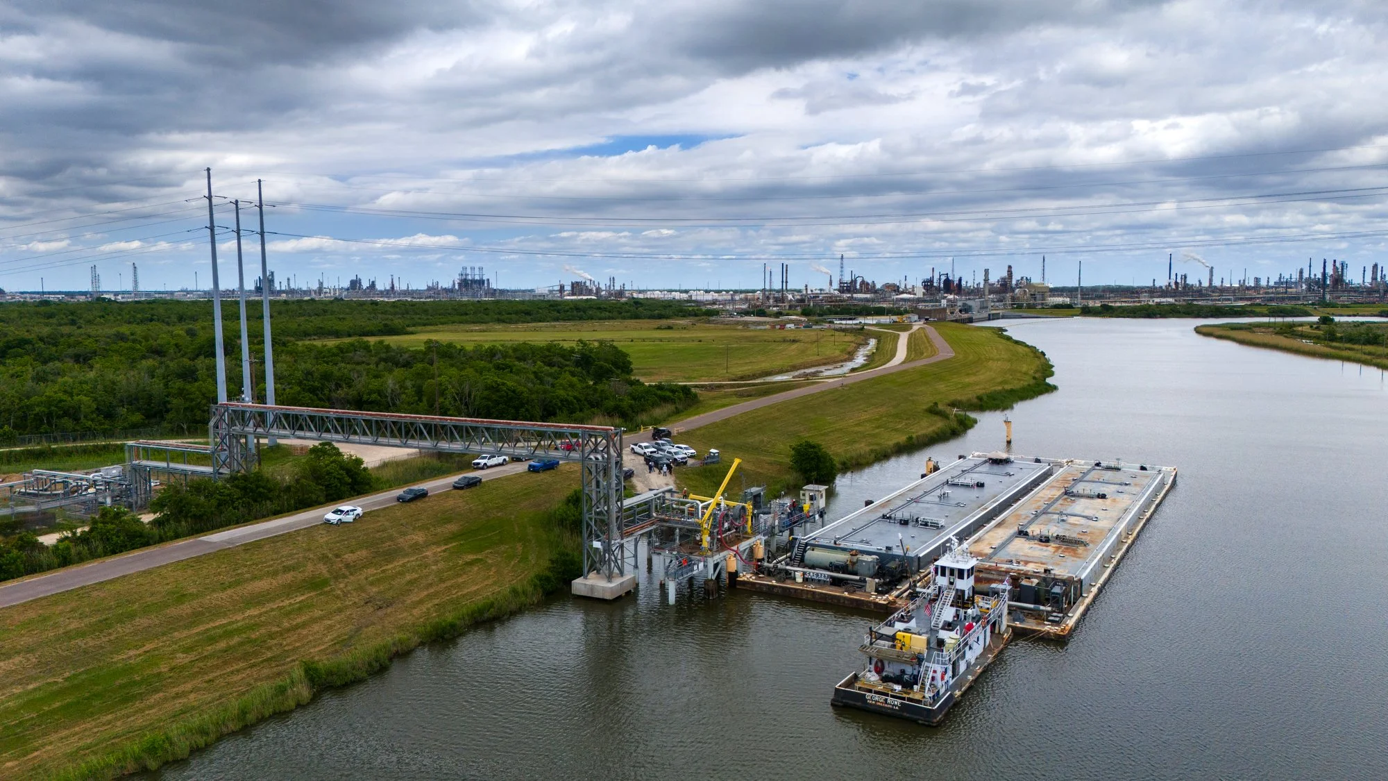 Industrial facility with a large barge on water, connected to a dock with equipment, green fields, trees, and a city skyline with factories and smokestacks in the distance under a cloudy sky.