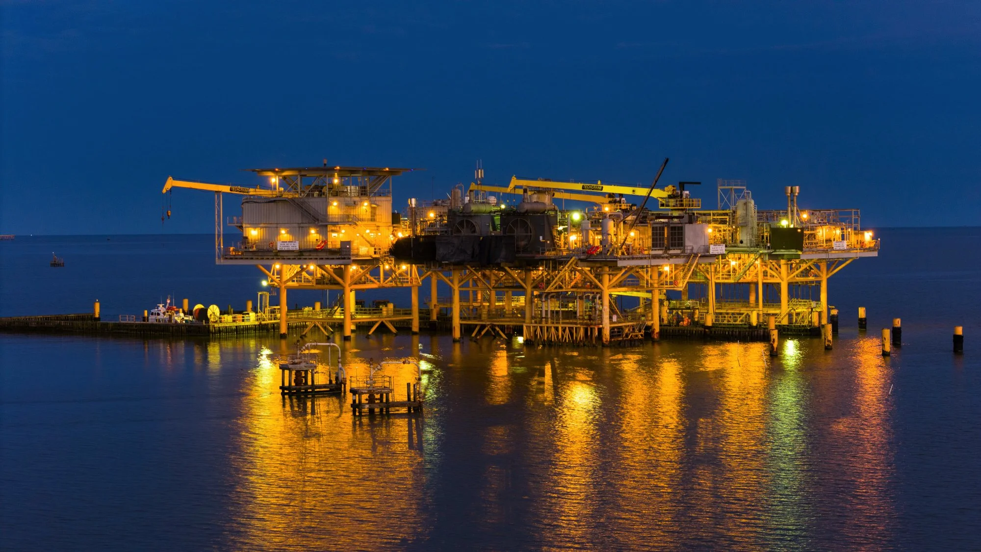 A brightly lit offshore oil rig at night reflecting in calm ocean waters.