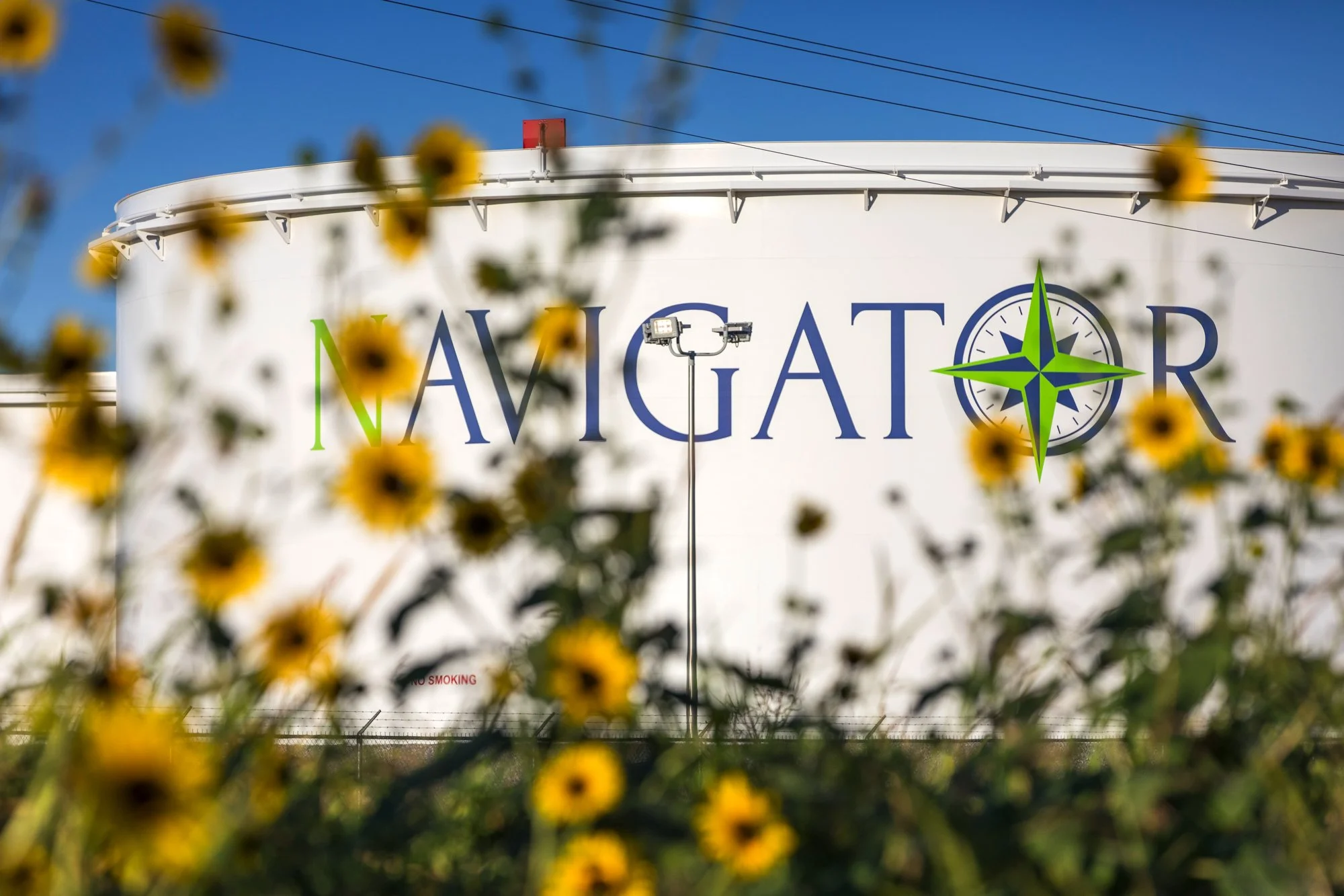 Large white tank with the word 'NAVIGATOR' and a green and blue compass logo, surrounded by yellow flowers and green foliage.