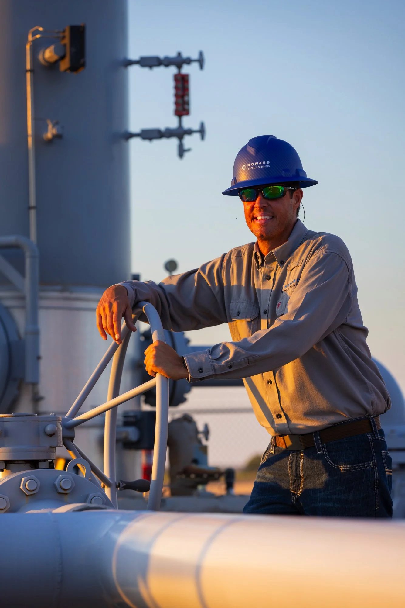 A man wearing protective sunglasses, a safety helmet, and a brown work shirt leaning on pipeline valve equipment outdoors during sunset.