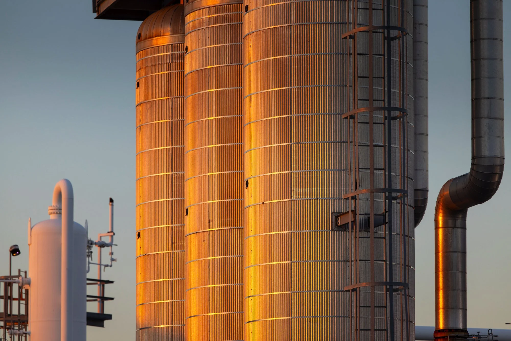 Close-up view of a modern industrial building with large, rust-colored metal silos and pipes during sunset.