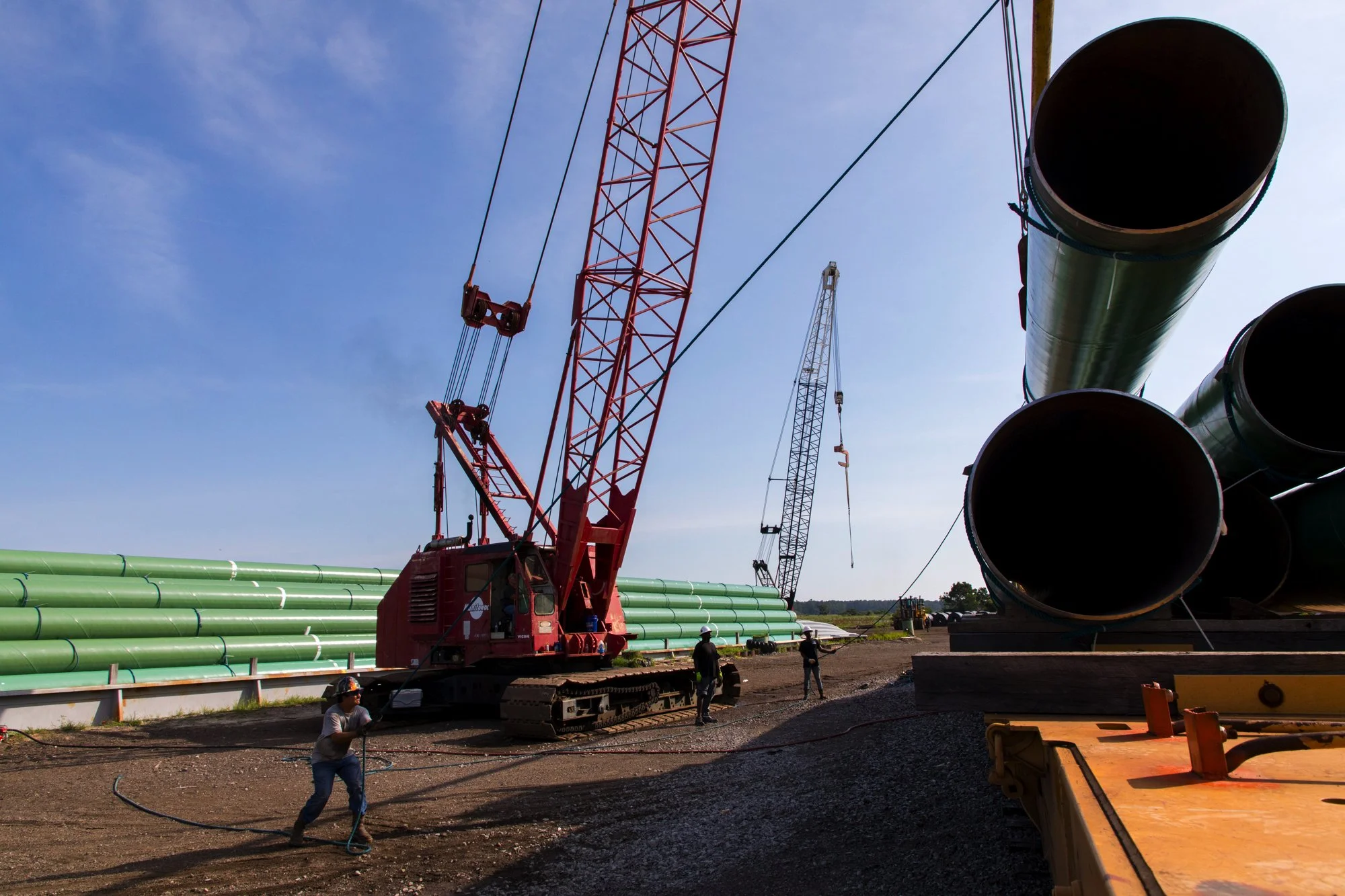 Worker holding a hose near construction equipment with large green pipes and cranes at a construction site.