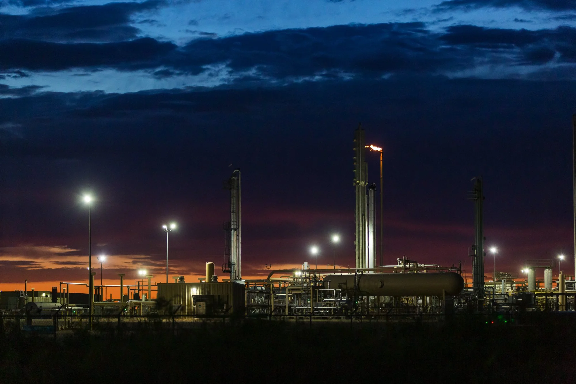 An industrial oil refinery at sunset with tall distillation towers, pipelines, storage tanks, and bright lights illuminating the facility against a darkening sky.