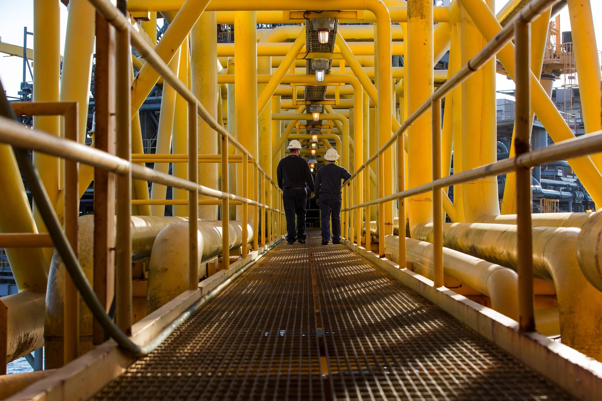 Two workers in hard hats walking on a metal catwalk among yellow industrial pipes and structures at an oil or gas facility.
