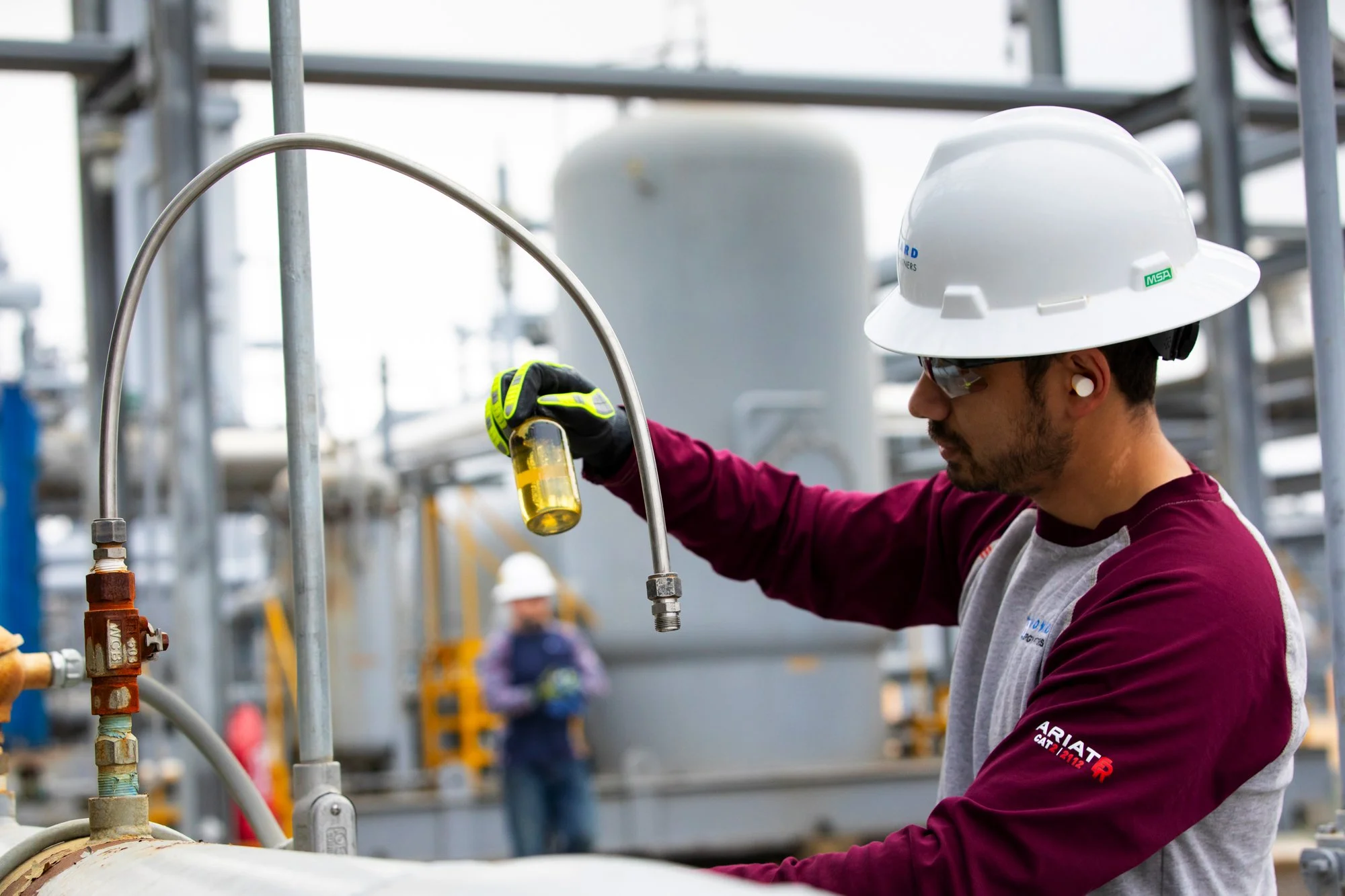 A worker with safety glasses, a white helmet, and ear protection working with industrial equipment outdoors, holding a yellow liquid container.