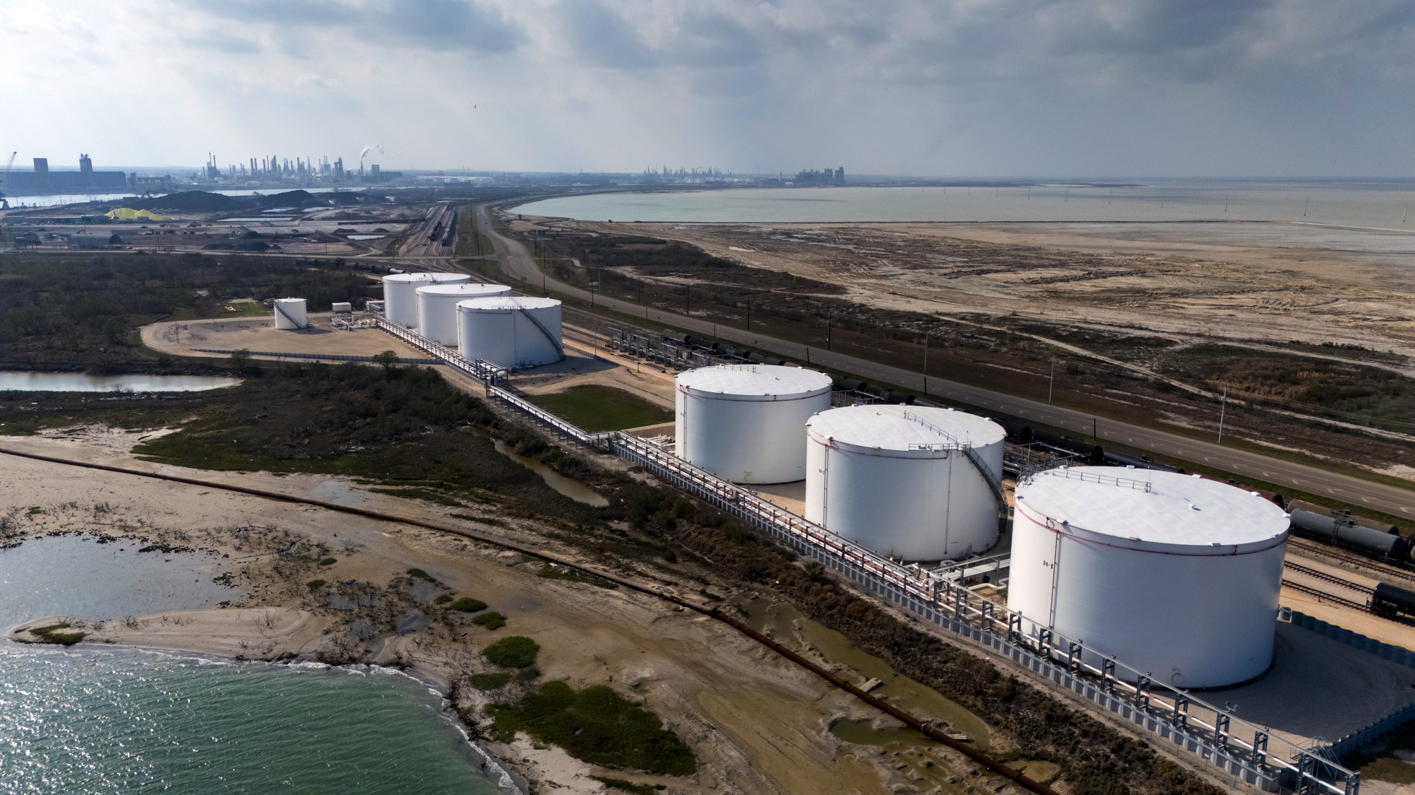 Aerial view of large white cylindrical tanks near a coast, with an industrial complex and oil refinery in the background, and a body of water in the foreground.
