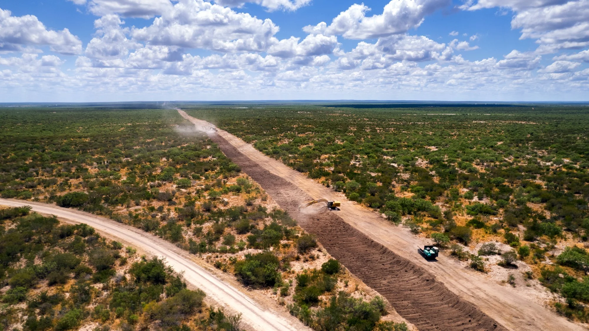 Aerial view of construction crews laying down pipeline in a vast, arid landscape with sparse vegetation and a partly cloudy sky.