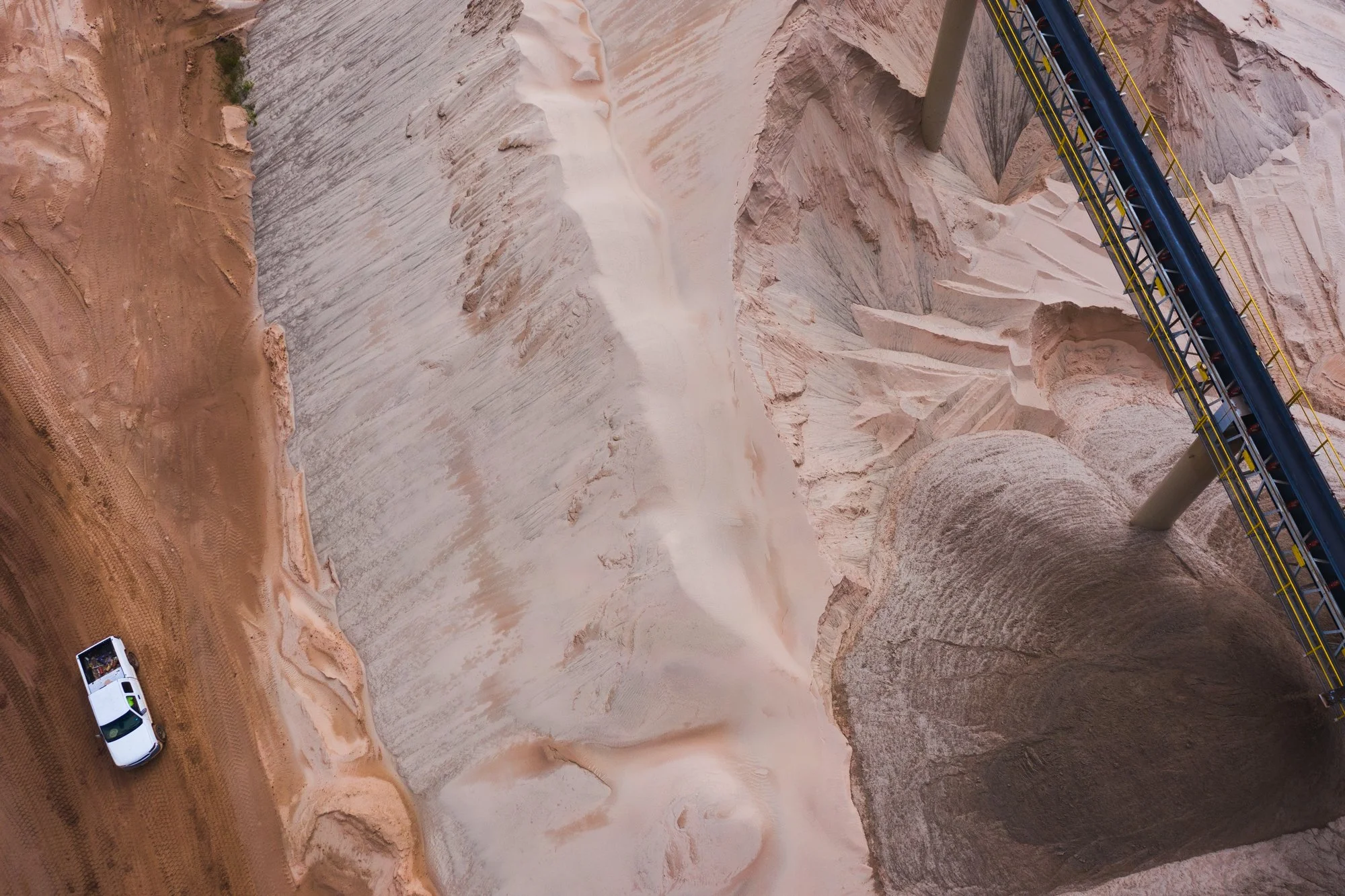 Aerial view of a large open-pit quarry with a conveyor system and a white pickup truck on dirt ground.