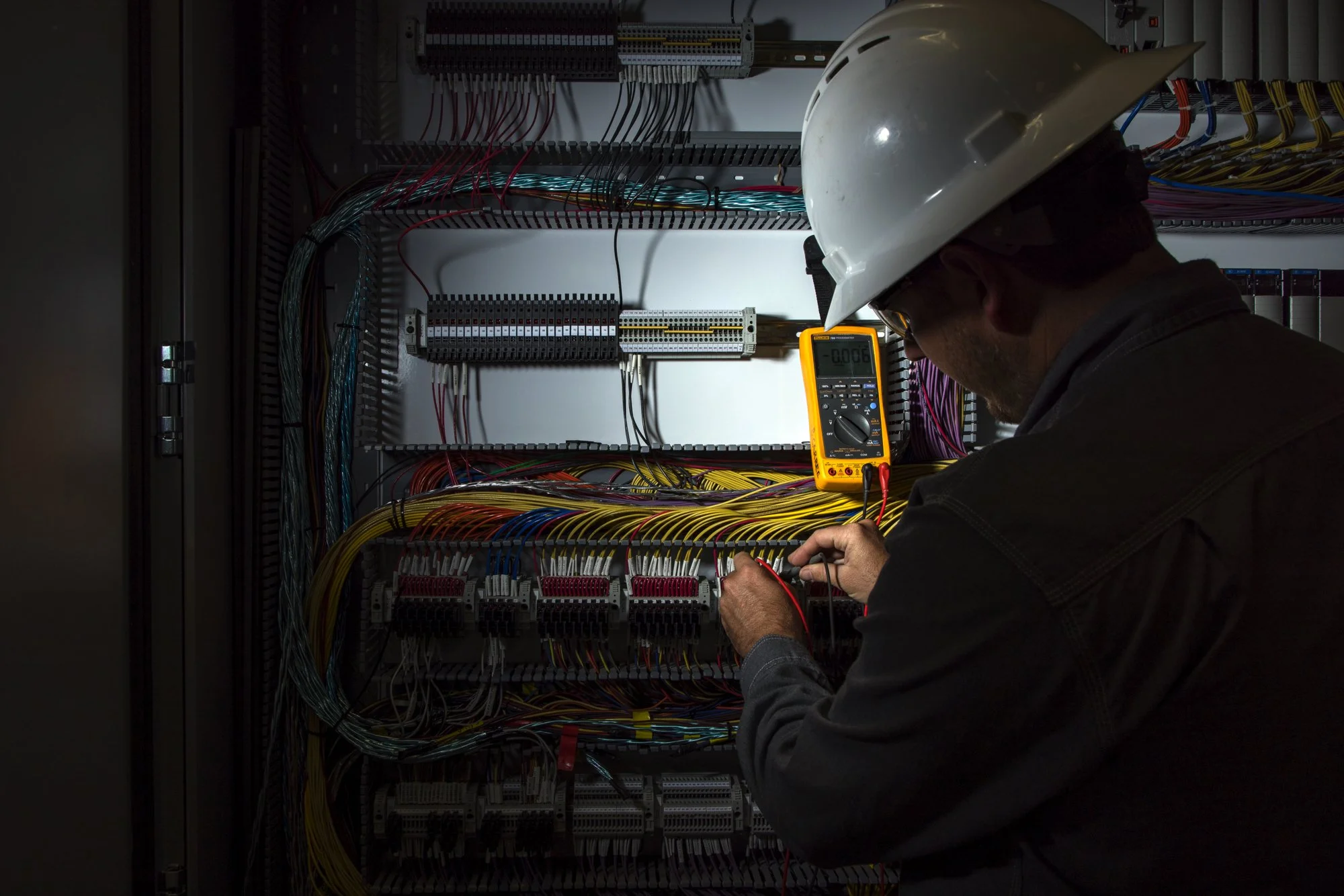 An electrician wearing a white safety helmet working on an electrical control panel with colored wires and using a digital multimeter.