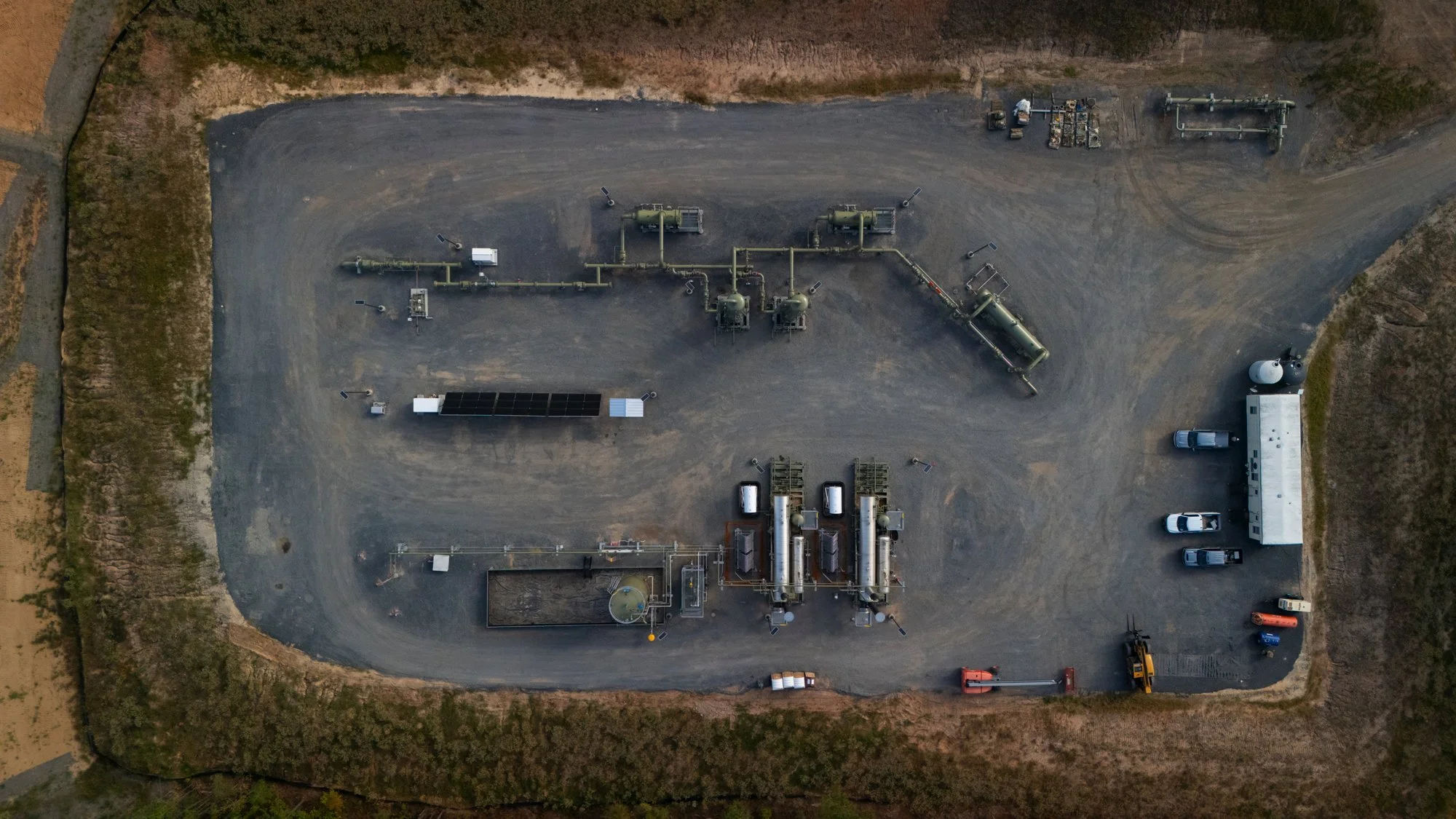 Aerial view of an industrial facility with pipelines, storage tanks, trucks, parked cars, and various equipment on a gravel surface surrounded by dirt and grass.