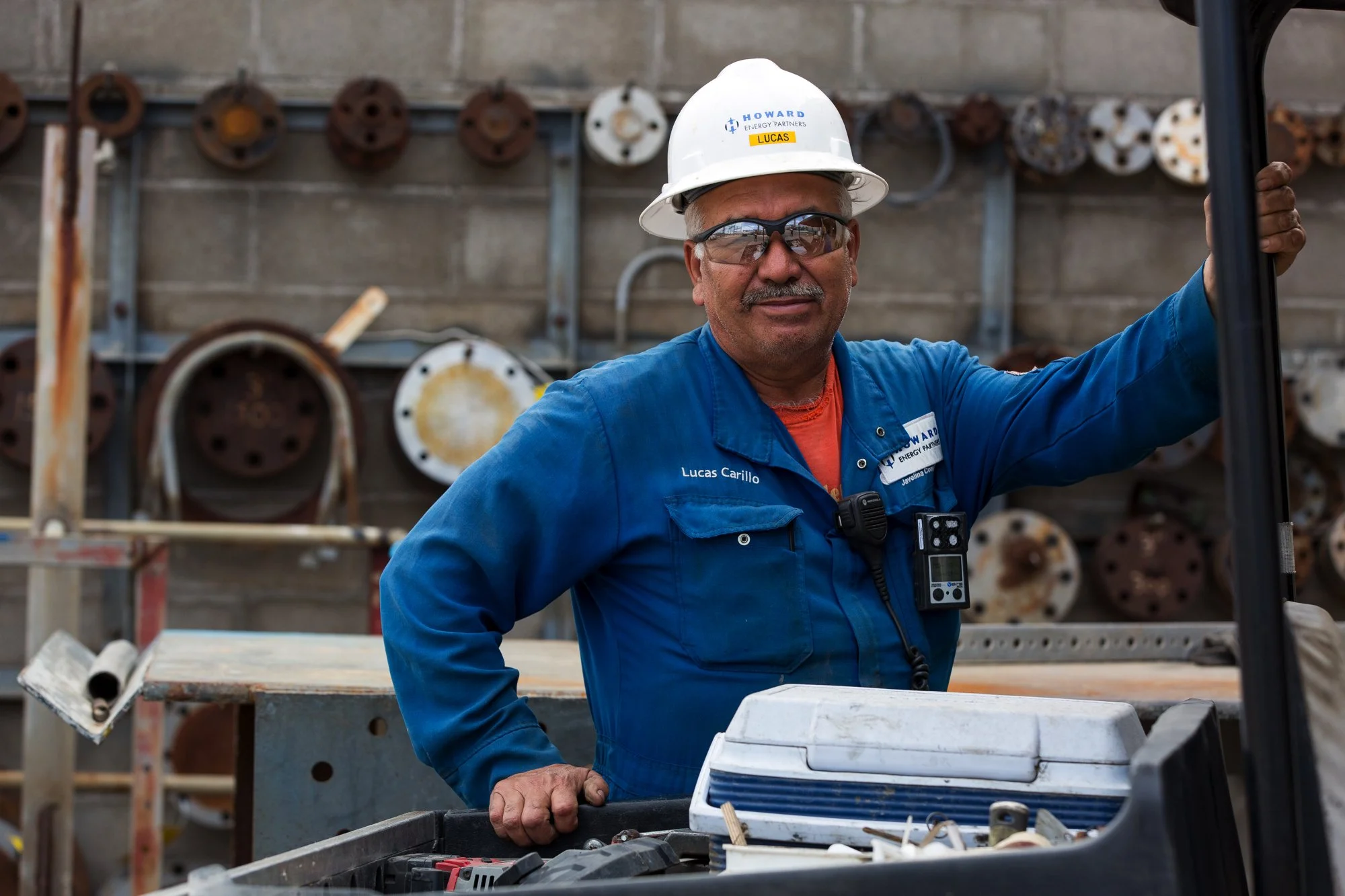 A man wearing safety glasses, a white safety helmet with the logo and the name tag 'Lucas' and a blue industrial jumpsuit, standing next to an open toolbox with tools inside, in a workshop or factory with rusty and clean pipes or machinery in the bac