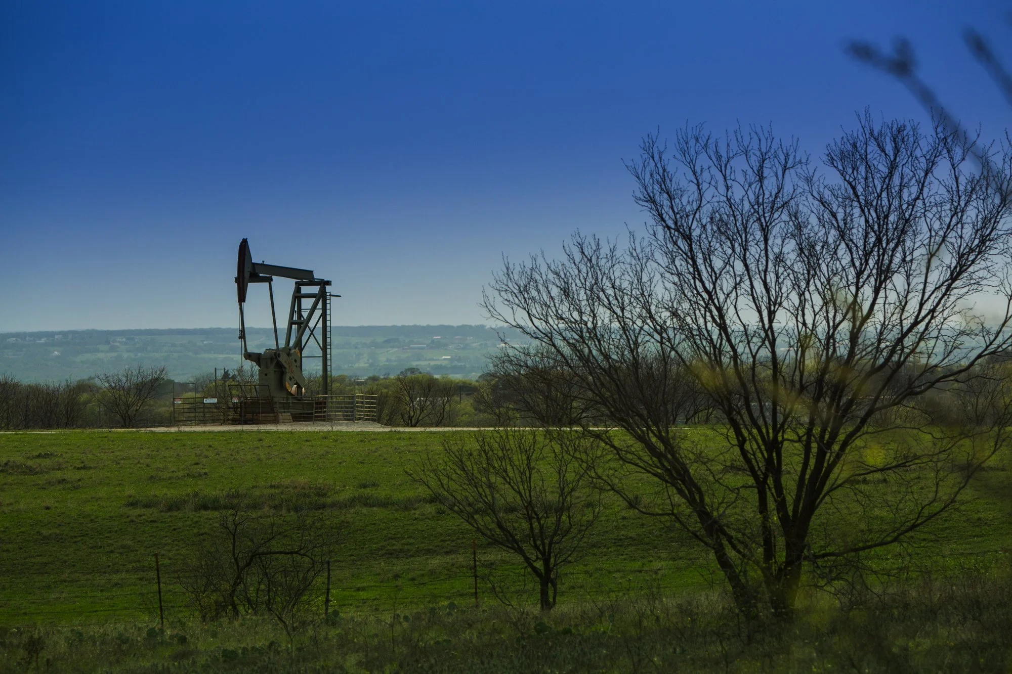 Oil pump jack in a grassy field with trees and hill in the background under a clear blue sky.