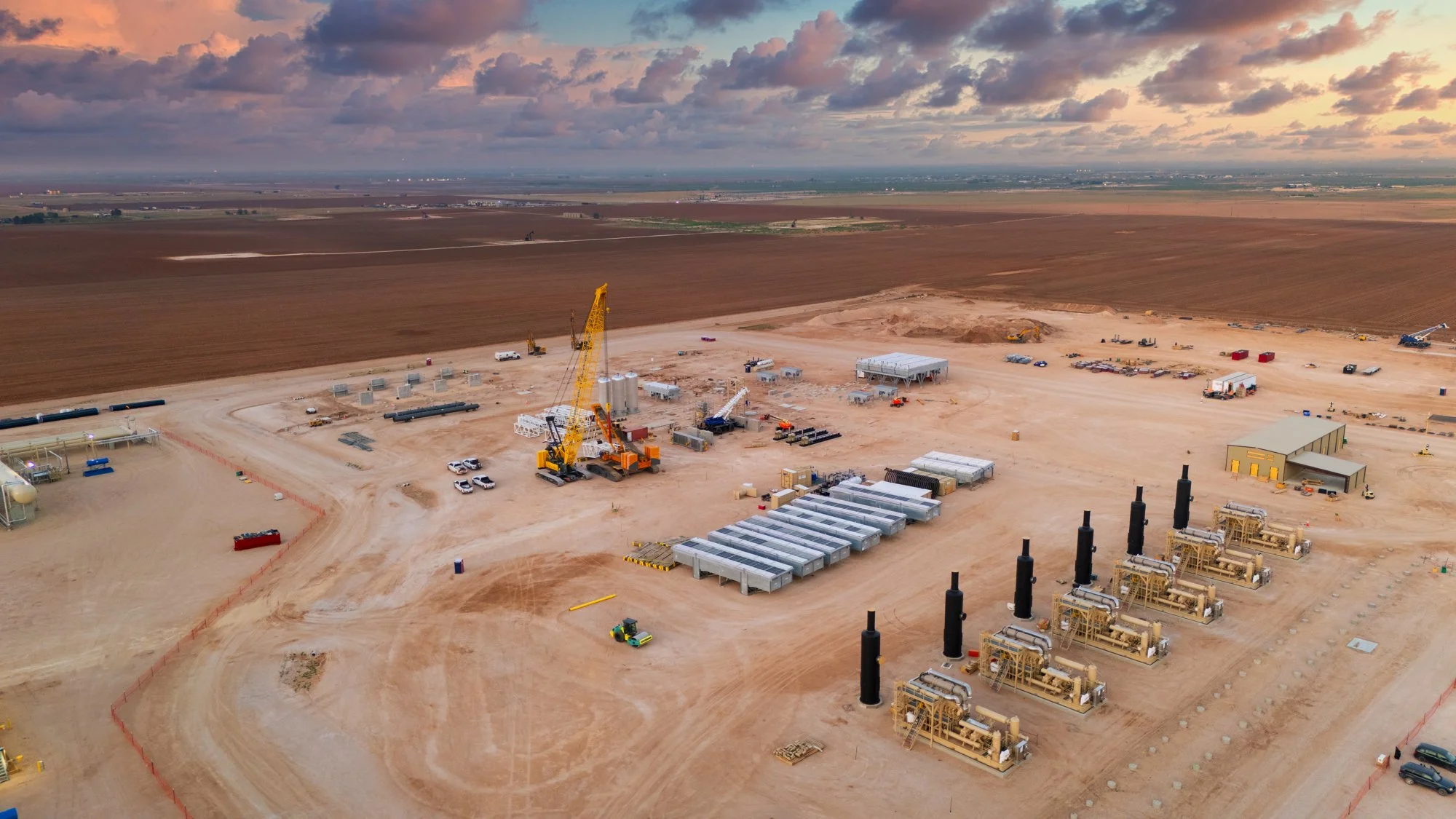 An aerial view of an oil or gas drilling site in a desert landscape with equipment, machinery, and vehicles, and open land extending into the horizon with clouds in the sky.