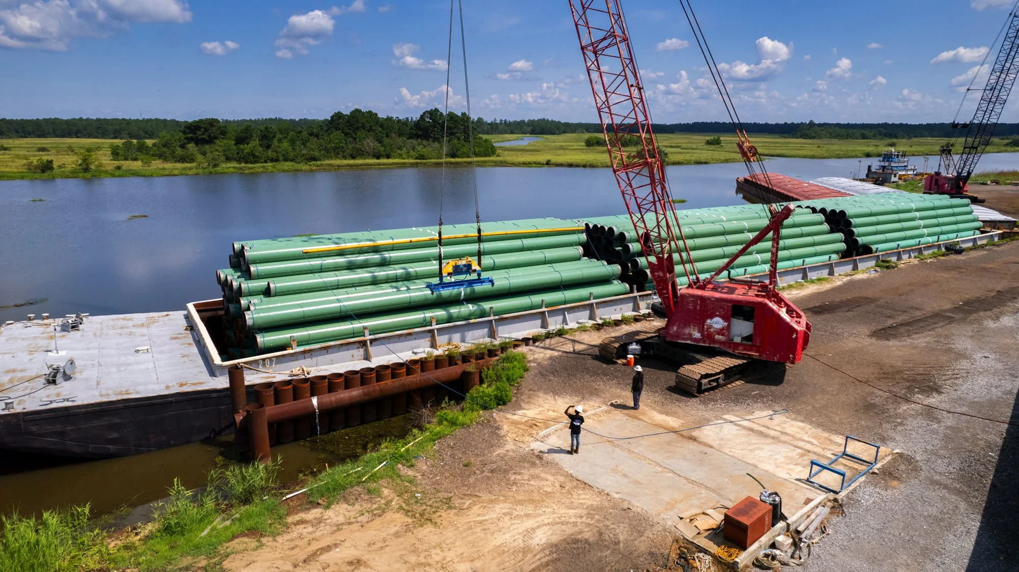 Large barge loaded with green pipes on a river, with a red crane lifting pipe sections, and two workers nearby, set in a flat landscape with water, trees, and a partly cloudy sky.