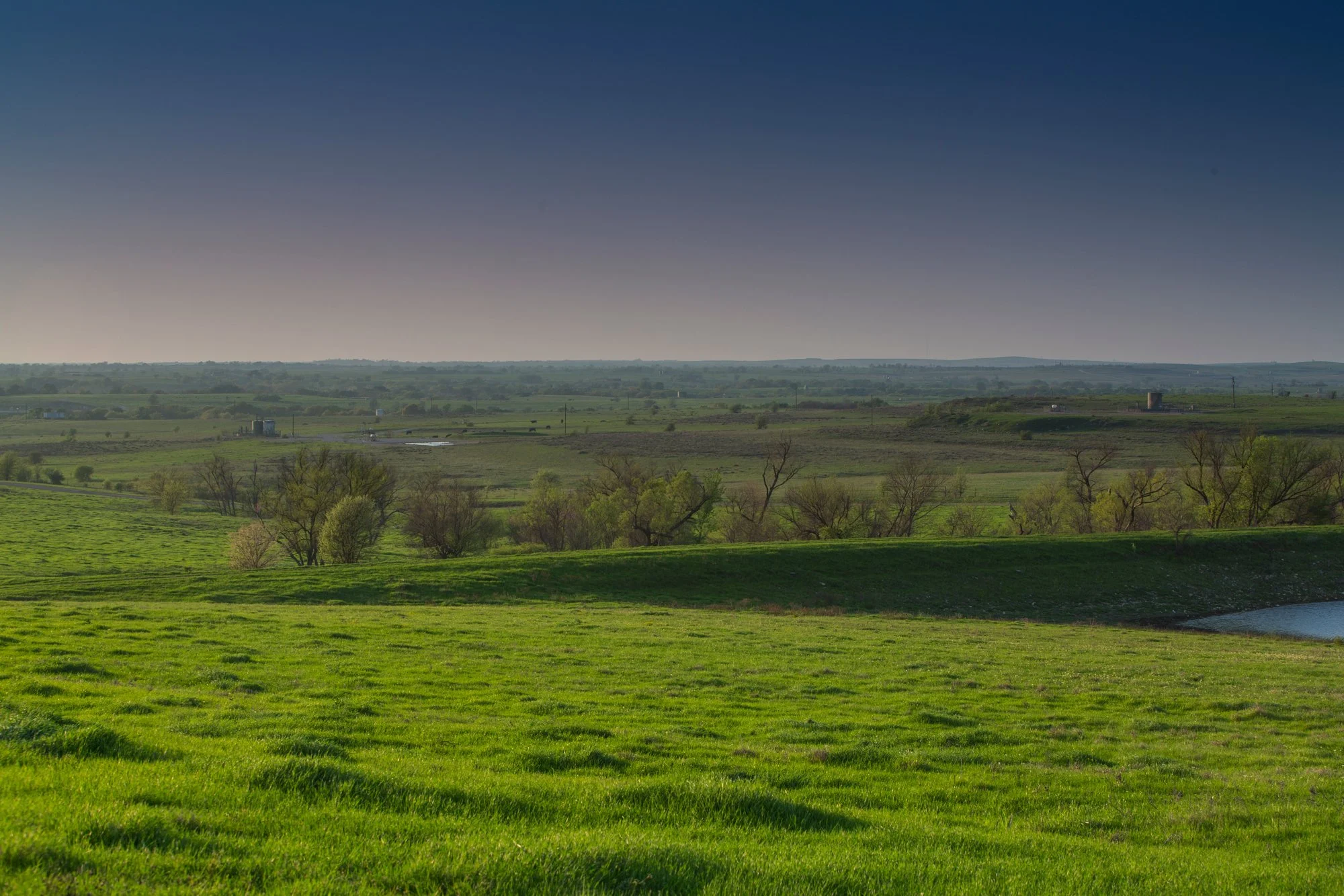 A wide landscape of green fields and trees with a small lake on the right, under a clear blue sky.
