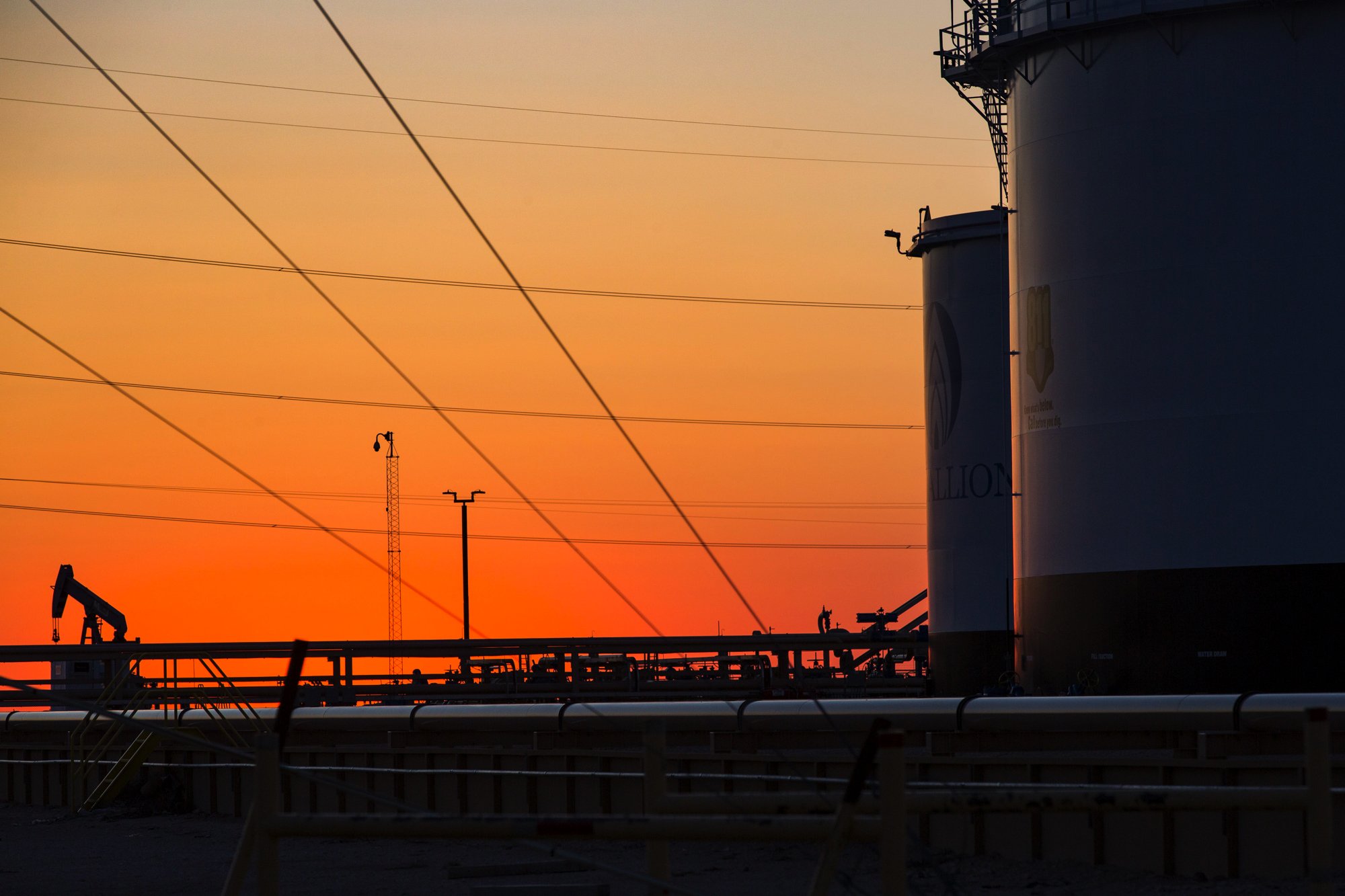 Silhouettes of oil tanks and oil pumps against a colorful sunset sky with power lines and utility poles in the background.