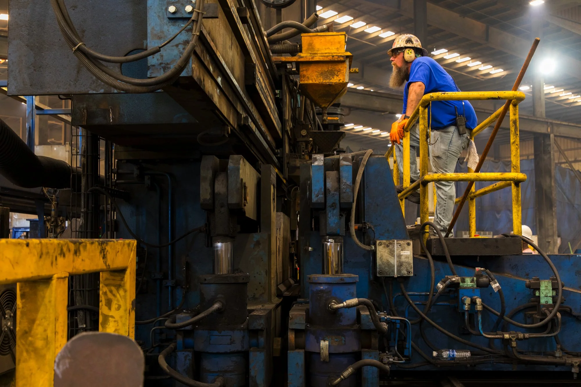 A worker in safety gear, including a helmet and ear protection, is standing on a yellow elevated platform, operating industrial machinery in a factory.