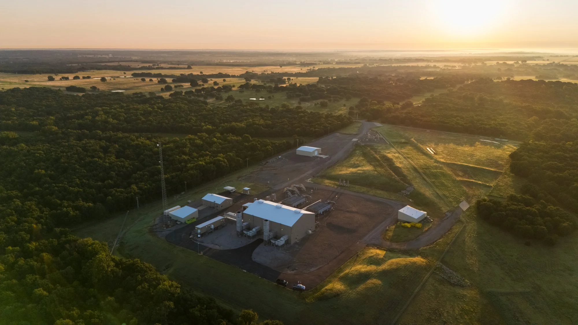 Aerial view of a rural industrial facility surrounded by green fields and trees at sunset, with several buildings, vehicles, and machinery visible.