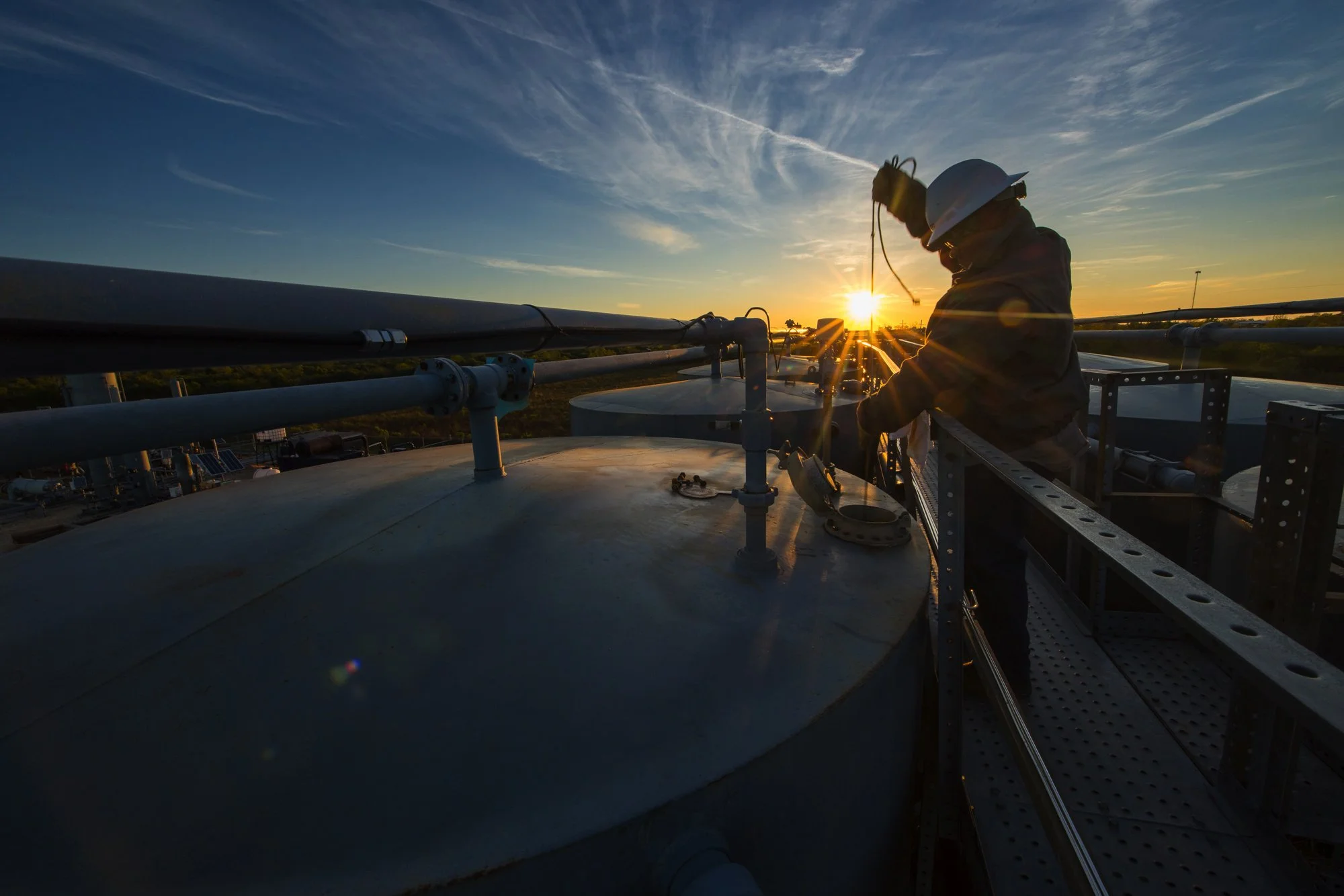 A worker wearing a hard hat and safety gear working on a large industrial tank at sunset, with piping and equipment around.