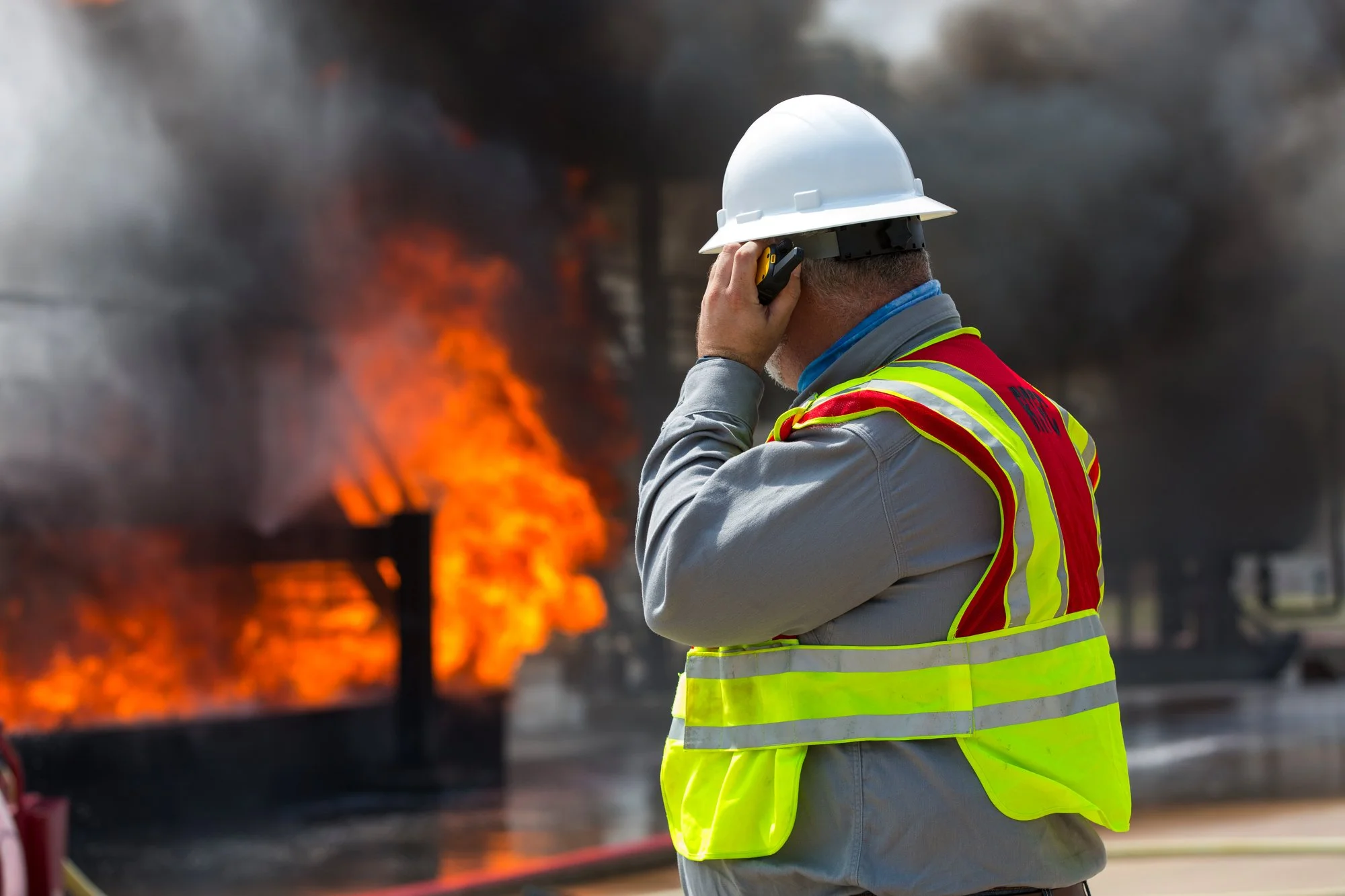 A firefighter in a gray uniform and yellow reflective vest wearing a white helmet, talking on a mobile phone while a building behind him is on fire with flames and black smoke.