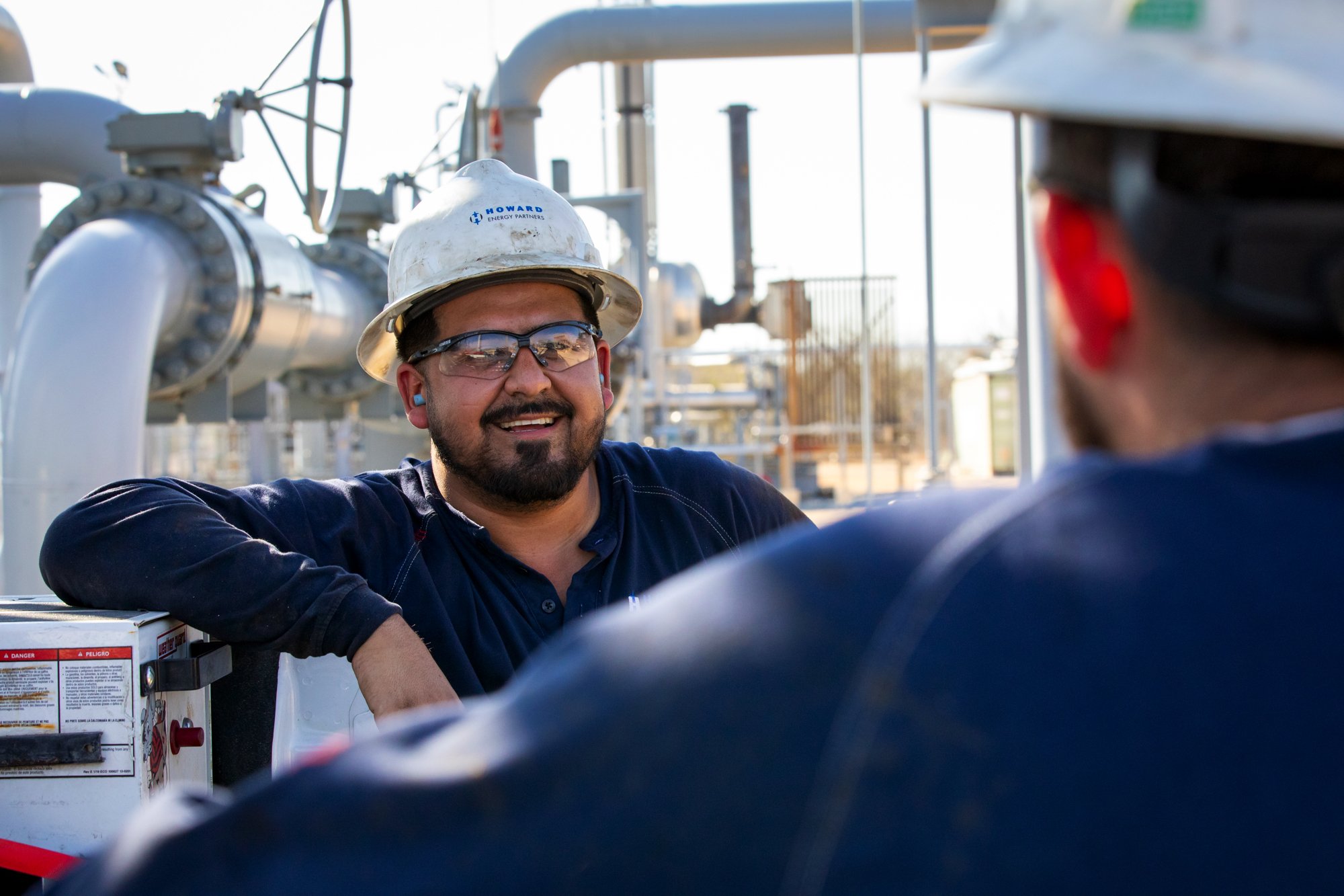 Two workers at an industrial site, one smiling and wearing safety glasses and a helmet, the other partially visible, both in safety gear, surrounded by piping and machinery.