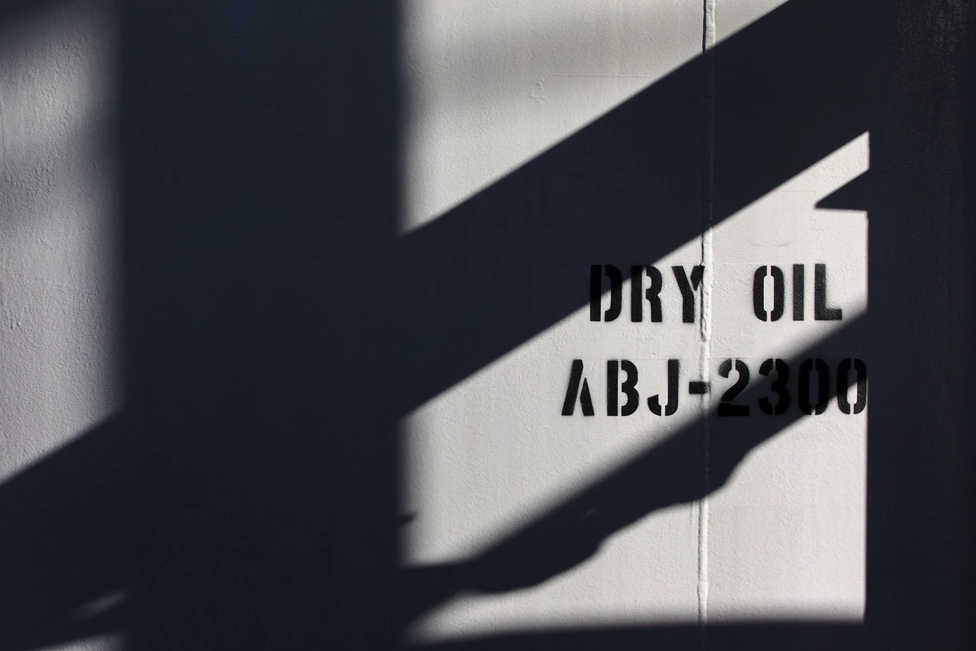 White wall with black stenciled text reading 'DRY OIL ABJ-2300' and shadows cast across it.