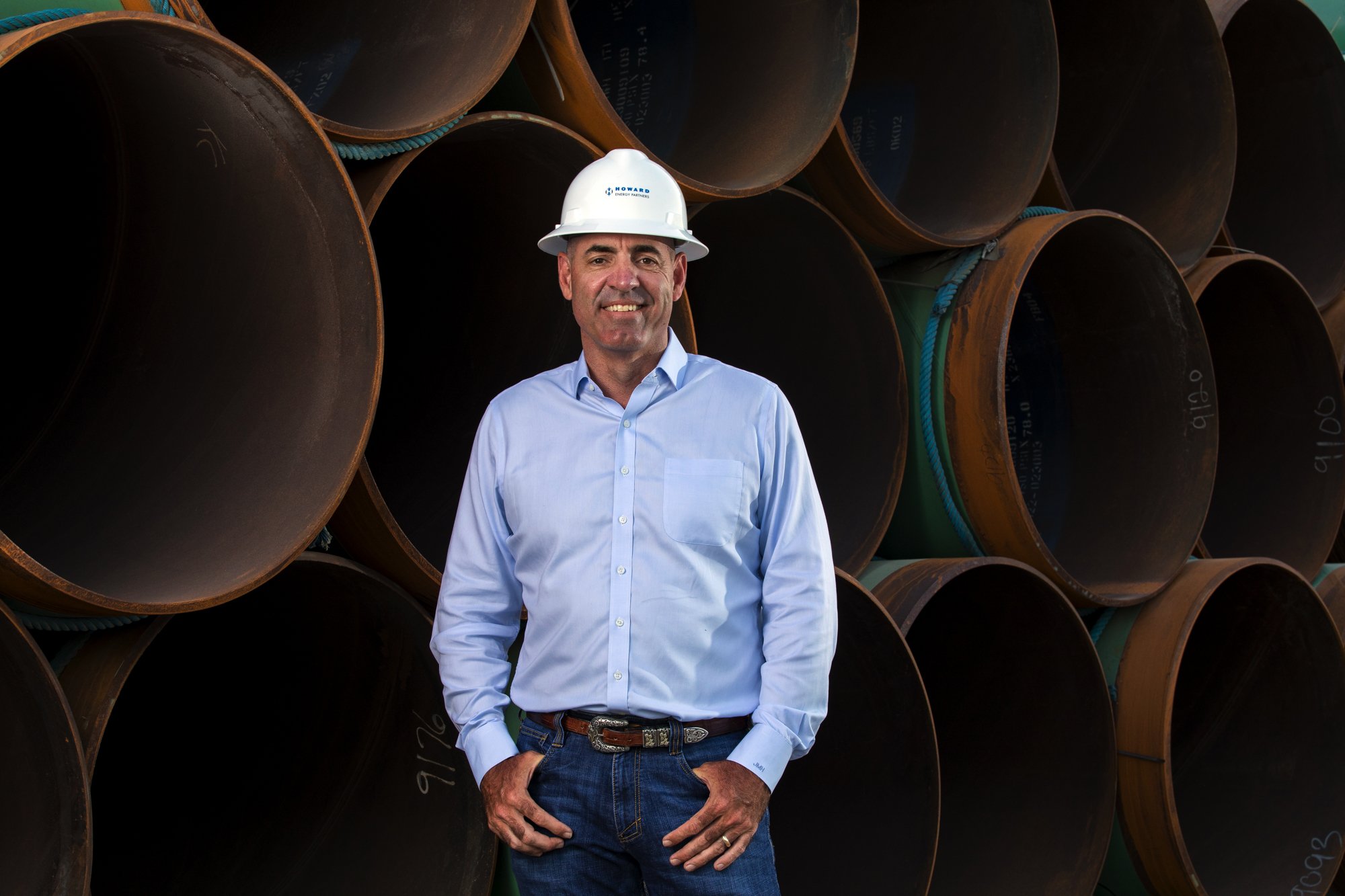 A man in a light blue shirt and jeans, wearing a white safety helmet, standing in front of stacked large industrial pipes.