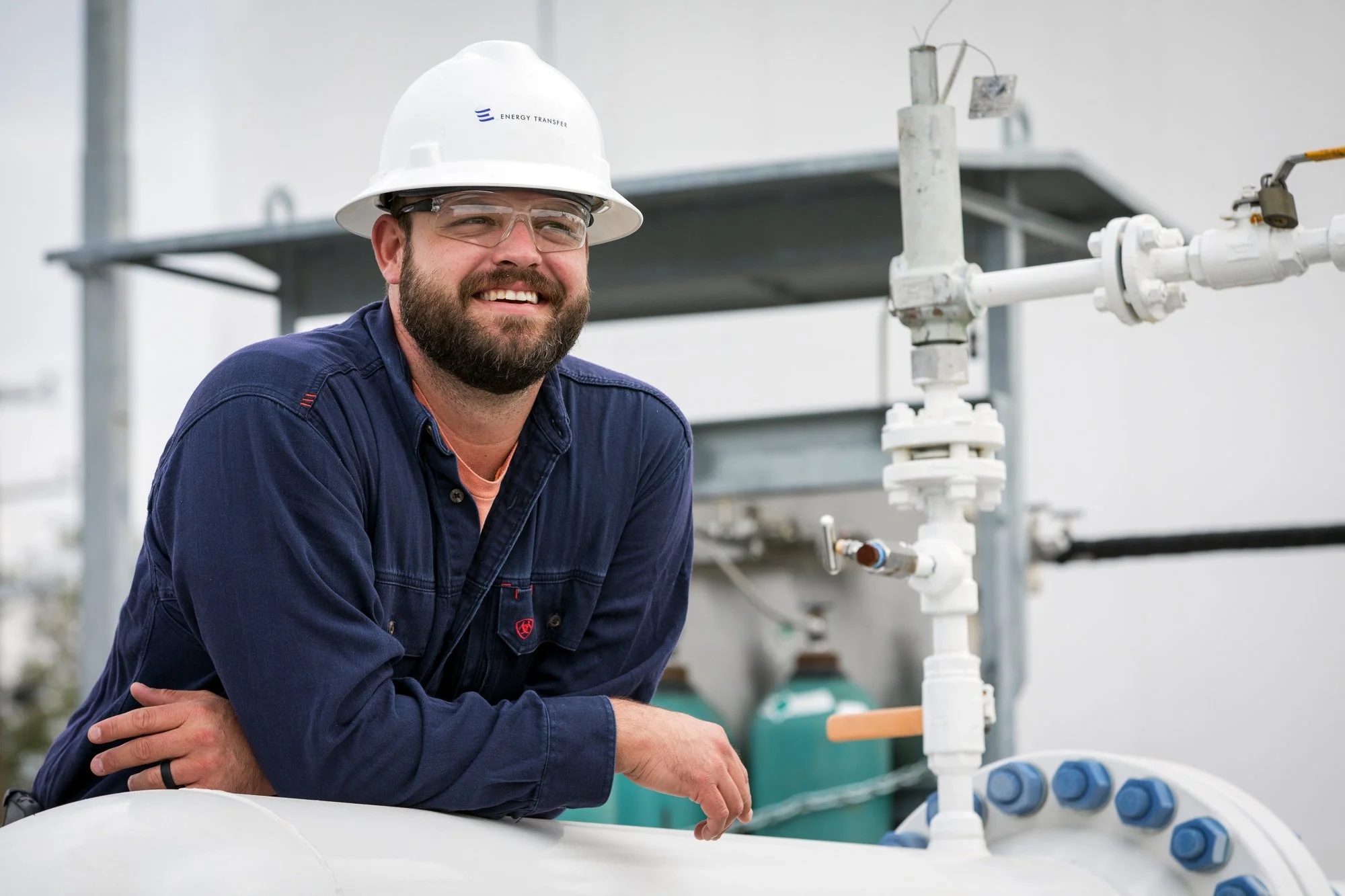 A man wearing safety glasses, a hard hat with the logo 'Energy Transfer,' and a navy blue work shirt is smiling and leaning on a large white pipe with blue bolts, in an industrial or mechanical setting.