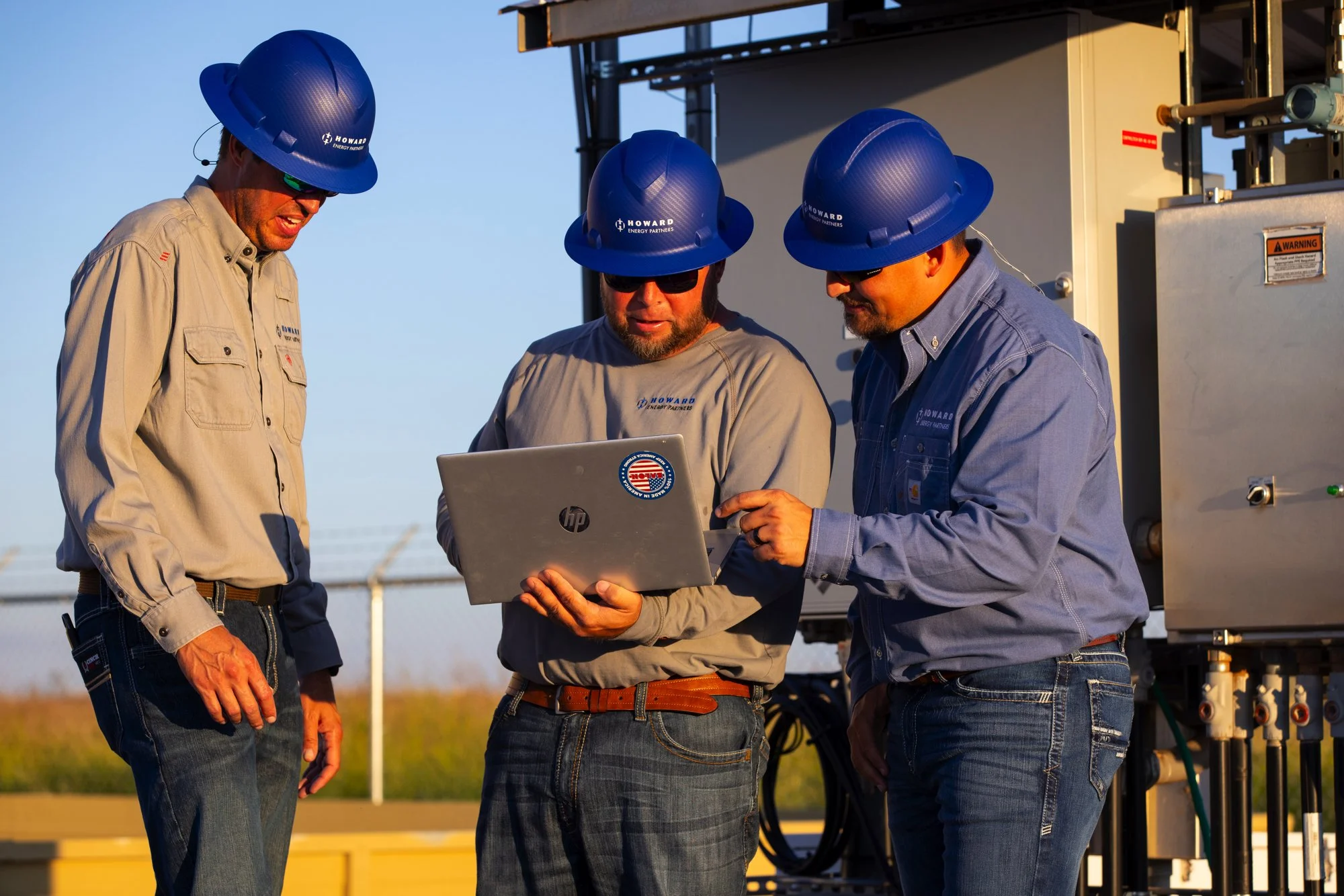 Three male workers wearing blue hard hats and safety glasses stand outdoors, looking at a laptop. They are dressed in work shirts with company logos, and there is industrial equipment behind them.