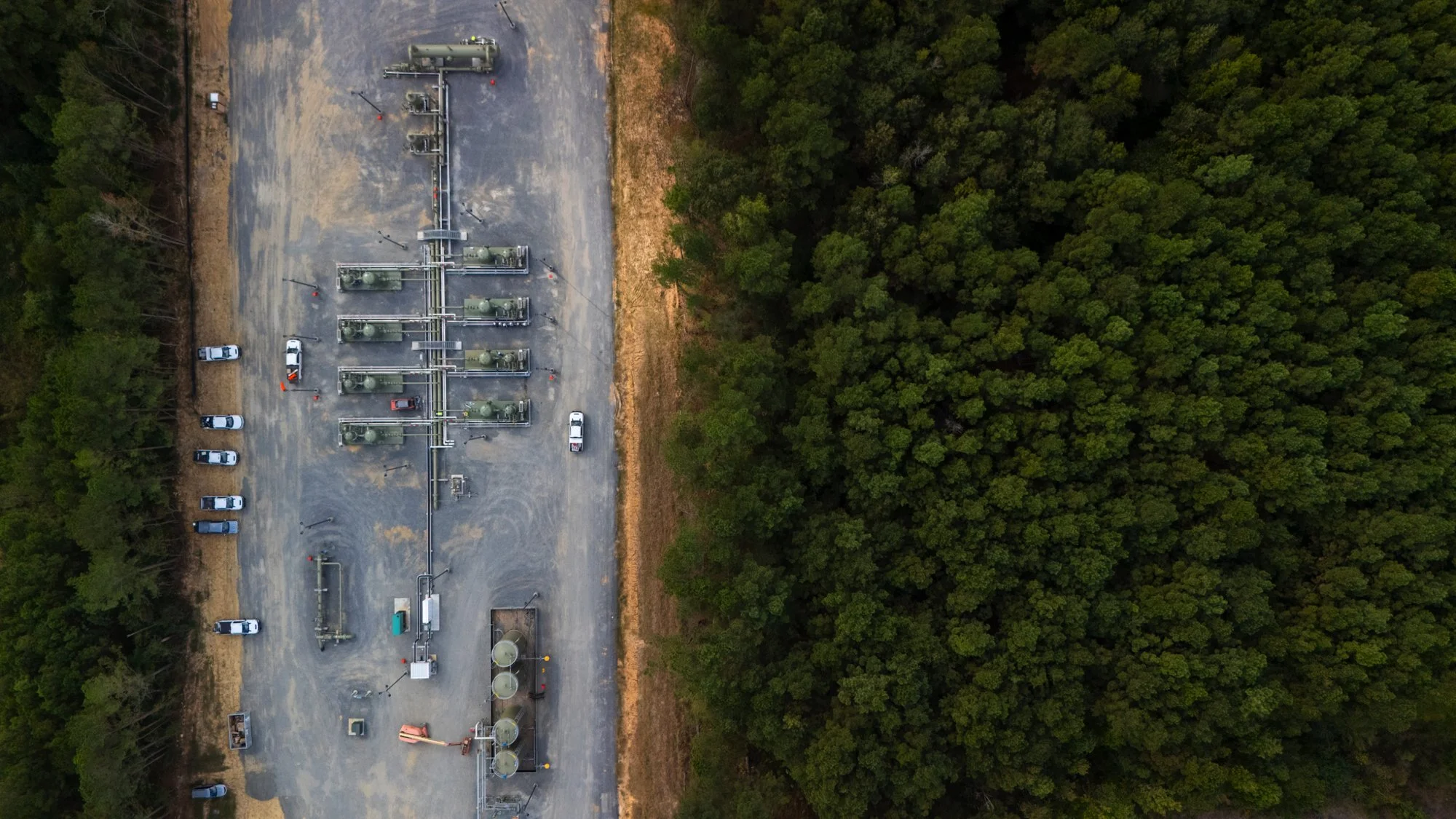 An aerial view shows a construction site and equipment on a gravel area, with a dense forest on the opposite side.