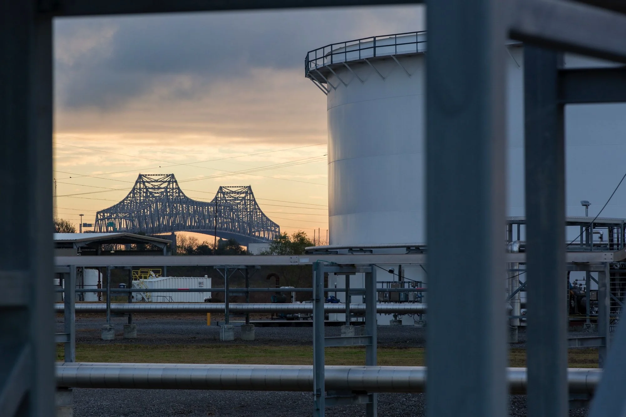 Industrial facility with storage tanks in the foreground and a bridge in the background at sunset.
