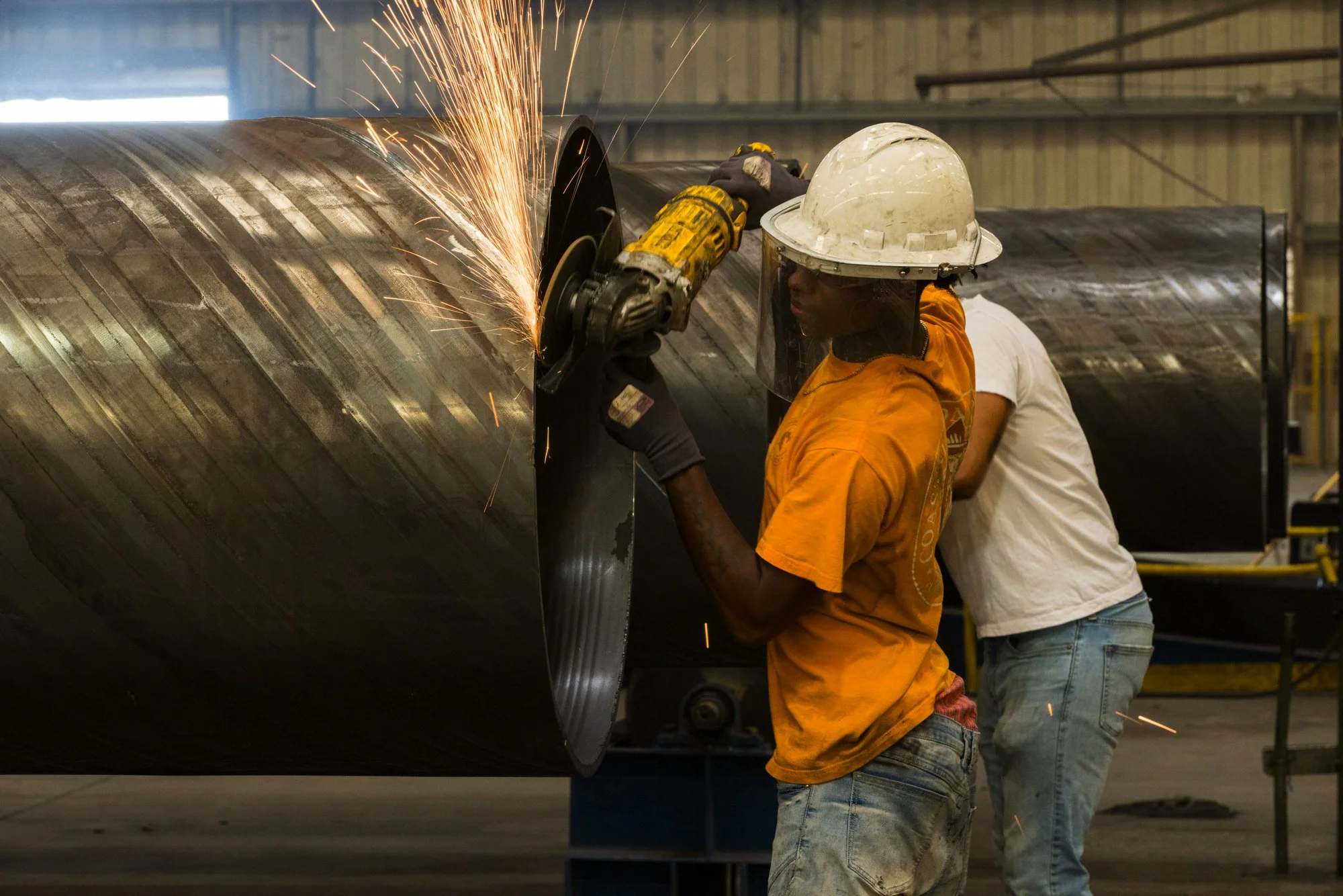 Two workers wearing hard hats and safety gear are using a grinding tool on a large metal pipe, producing sparks inside an industrial warehouse.