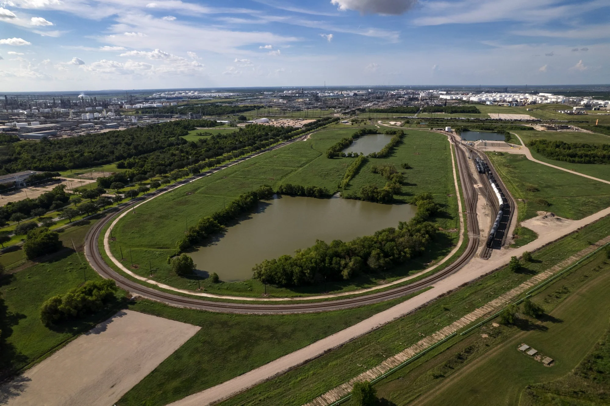 Aerial view of a park with a pond, a train on a curved track, green fields, and a distant industrial area under a partly cloudy sky.