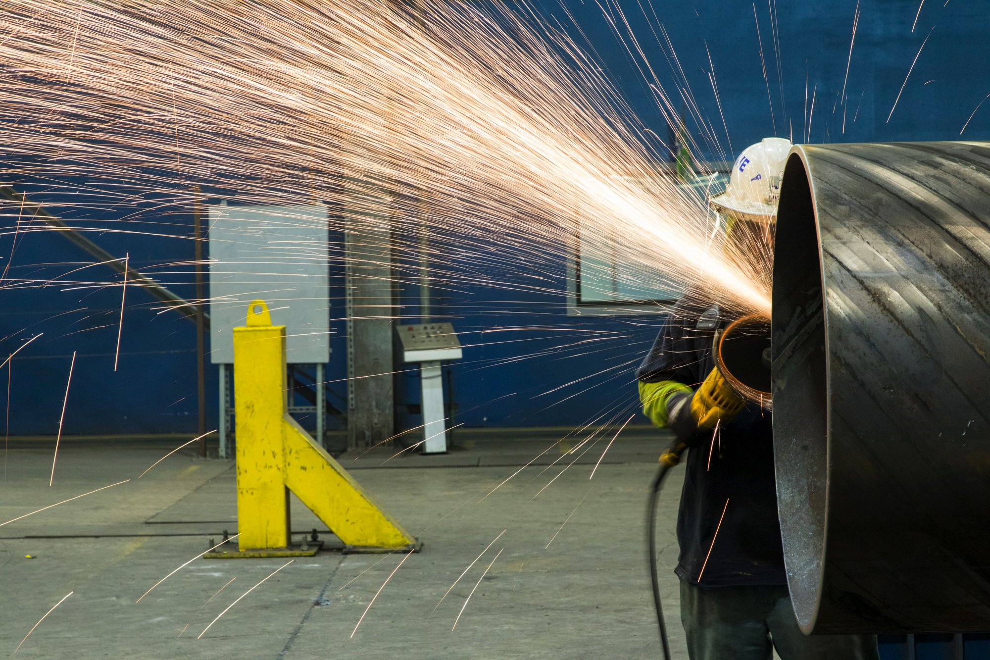 A worker wearing a helmet and safety gear welding a large metal pipe, producing sparks in an industrial setting.