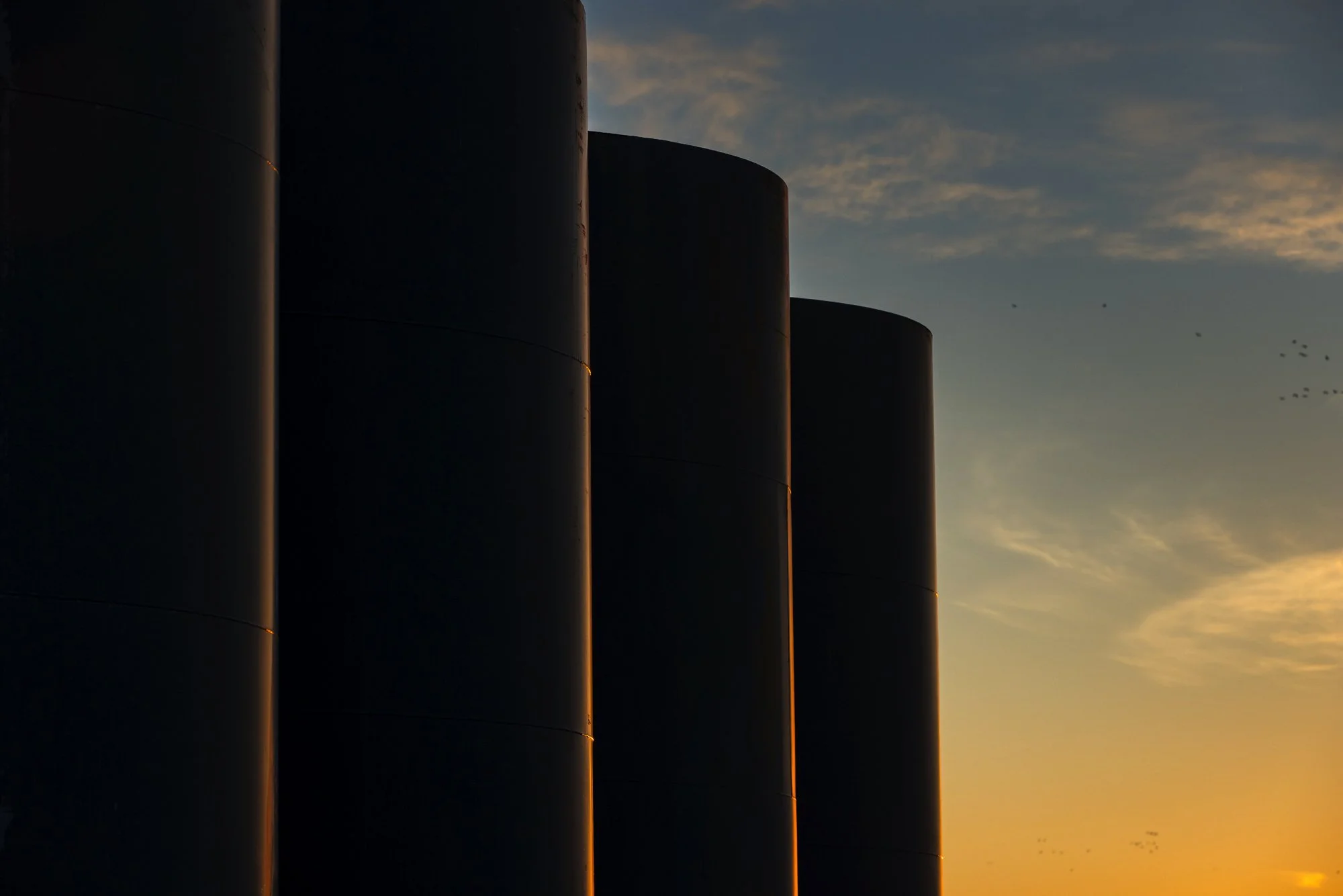 Close-up of dark metallic industrial tanks with a sunset sky in the background.
