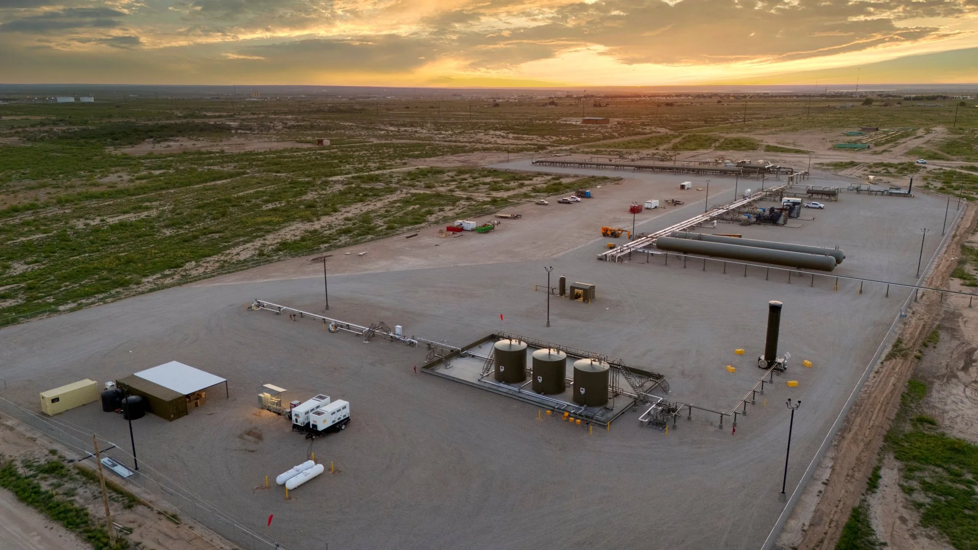 A large industrial site with pipelines, storage tanks, and vehicles in a desert landscape at sunset.